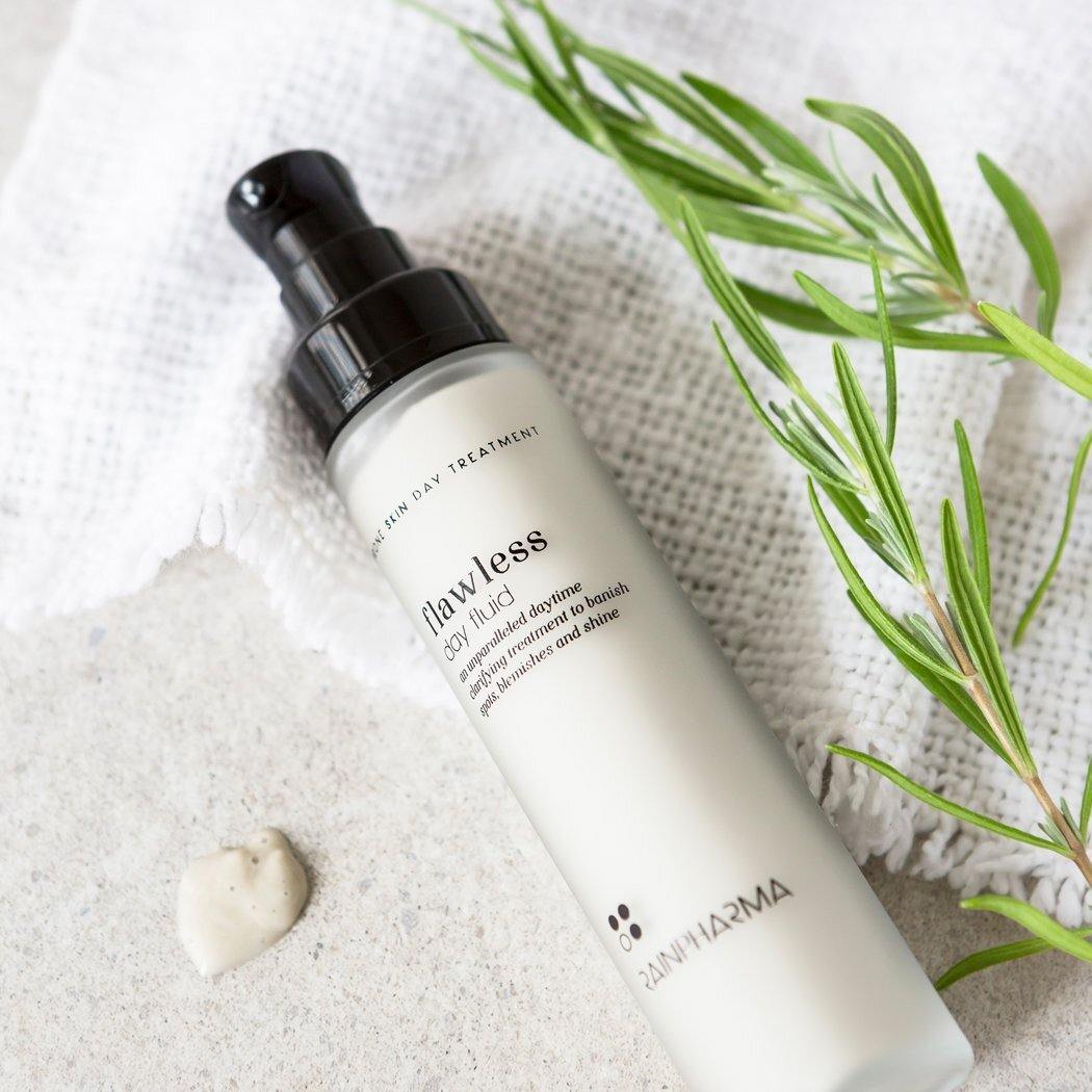 Een flesje RainPharma Flawless Day Fluid - Dagcrème onzuivere van vette huid rust op een wit gestructureerd doekje, naast een groen takje en een dotje crème op een licht oppervlak.