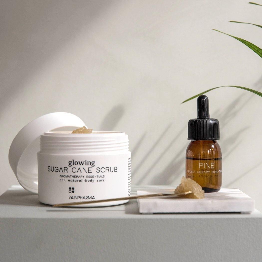 Een wit potje RainPharma Body Polish (Glowing Sugar Cane Scrub) met een open deksel en een bruin druppelflesje staan op een wit oppervlak, elk met een scrub doordrenkt met essentiële oliën op een spatel.
