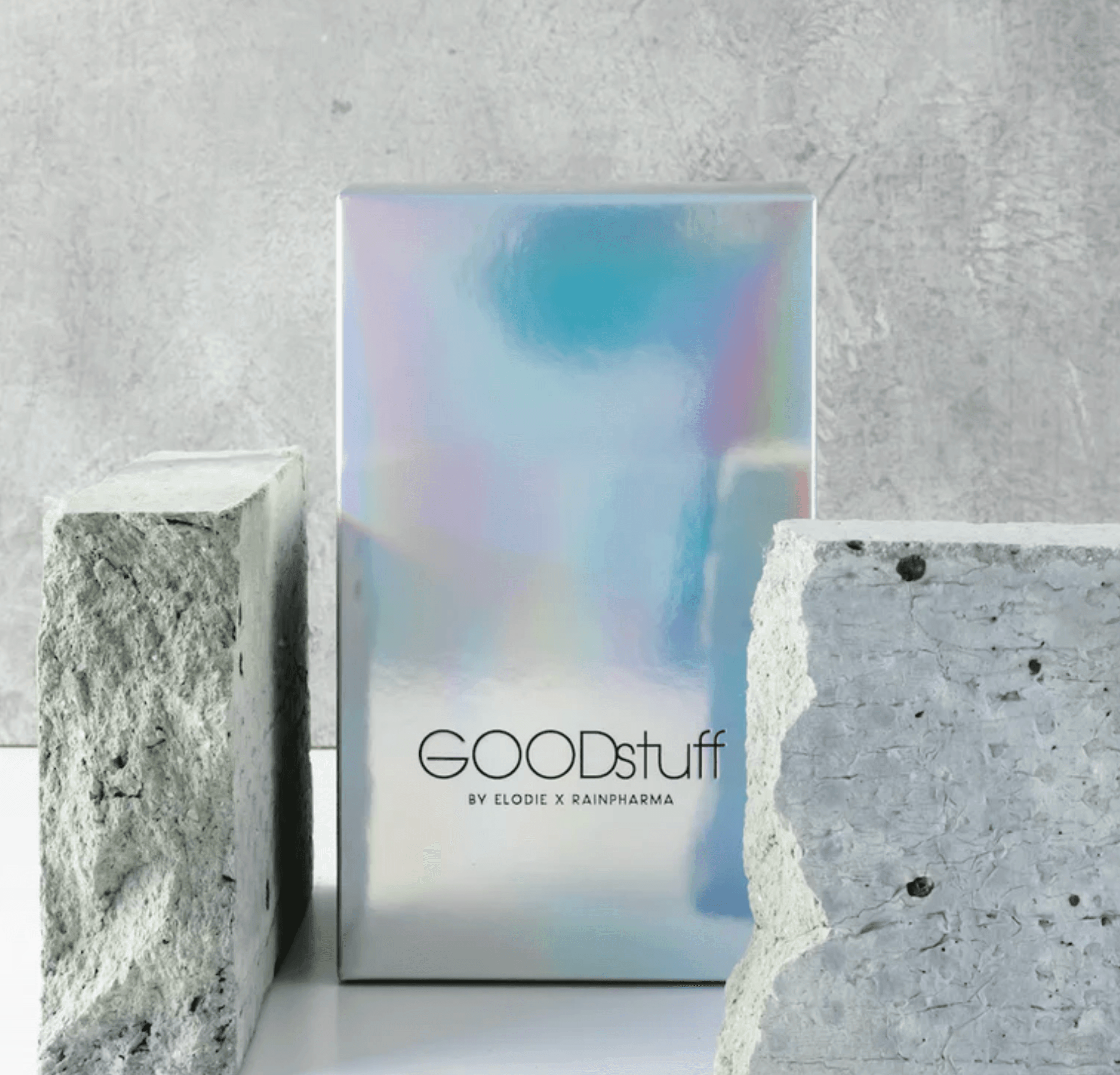 Een holografische doos met het label "Good Stuff by Elodie x RainPharma", gevuld met essentiële eiwitten, staat elegant tussen twee betonnen blokken op een grijs oppervlak.
