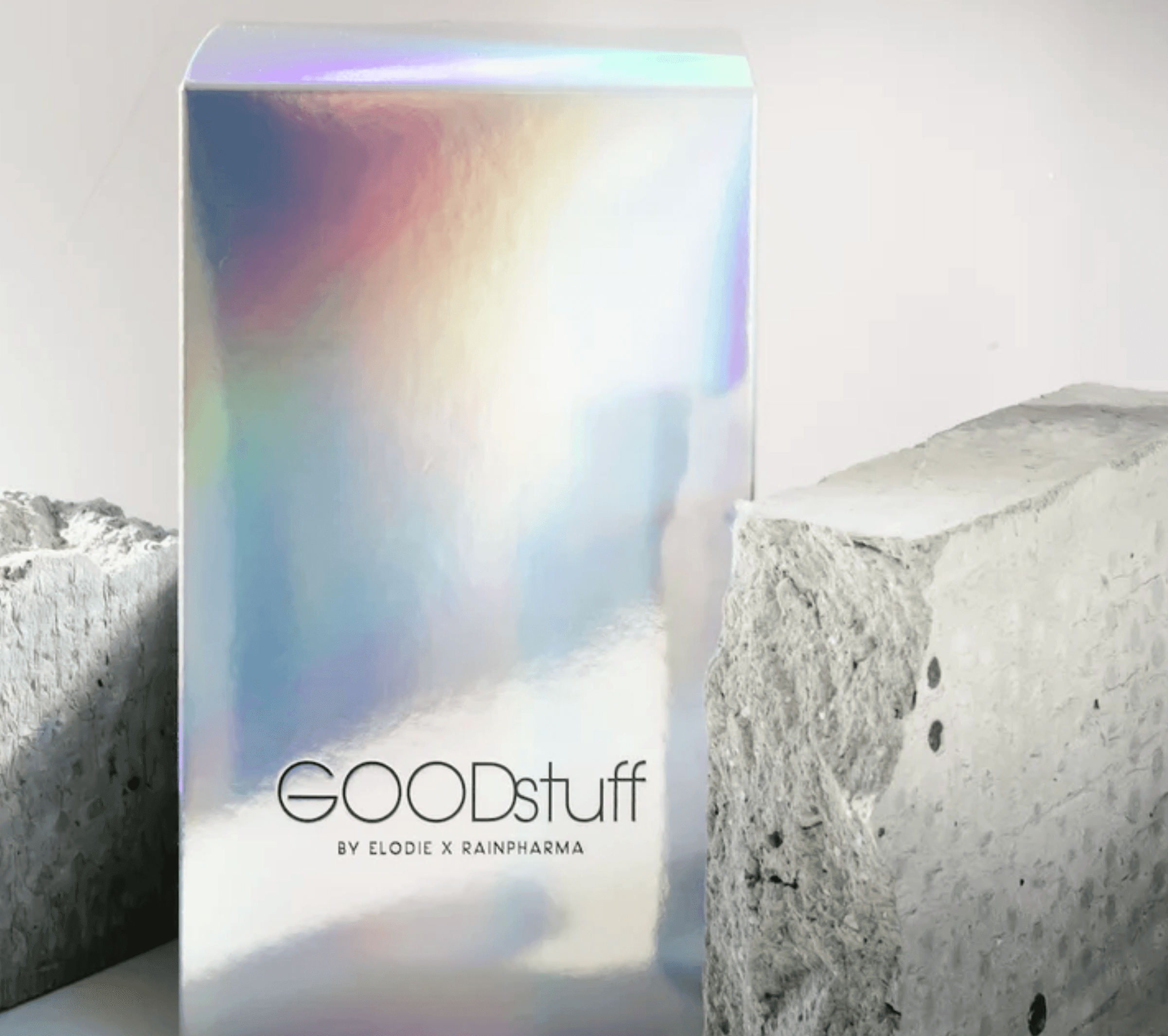 Een holografische doos met het label "Good Stuff by Elodie x Rainpharma" wordt samen met twee concrete stukken gepresenteerd, die een premium voedingssupplement aanbieden dat de opbouw van spiermassa ondersteunt met hoogwaardige eiwitten.