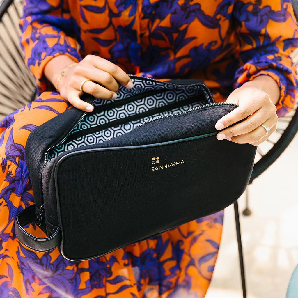 Iemand in een opvallende oranje en paarse jurk opent de RainPharma Toilettas - Maxi - Black, met een geometrische zwart-witte binnenkant, ideaal om verzorgingsproducten georganiseerd in op te bergen tijdens het reizen.