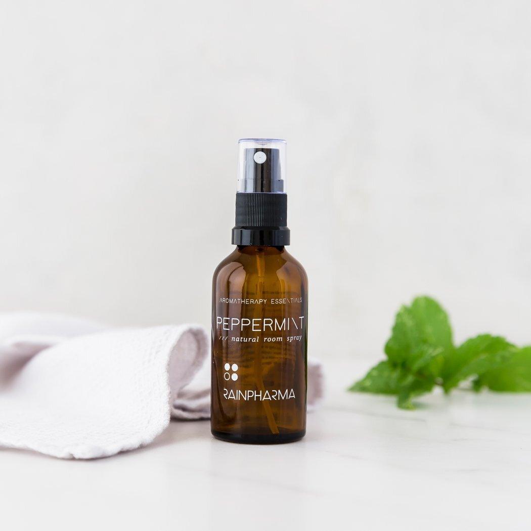 Een bruine glazen fles RainPharma Natural Room Spray Pepermunt staat op een wit oppervlak naast een gevouwen doek en verse muntblaadjes.