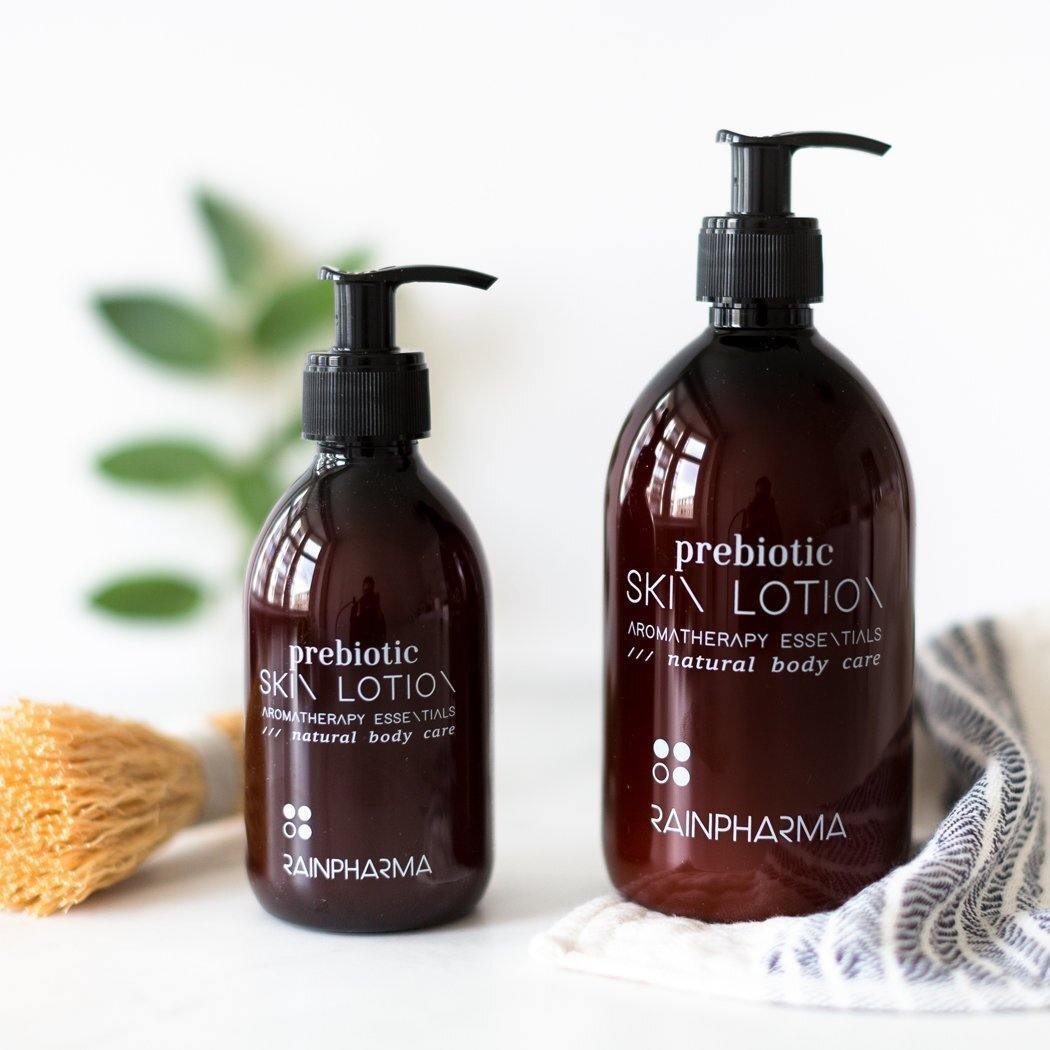 Twee bruine pompflacons van RainPharma Prebiotic Skin Lotion staan naast een natuurlijke spons en handdoek op een wit oppervlak en benadrukken de focus van het merk op plantaardige huidverzorging en zachte prebiotische formules.