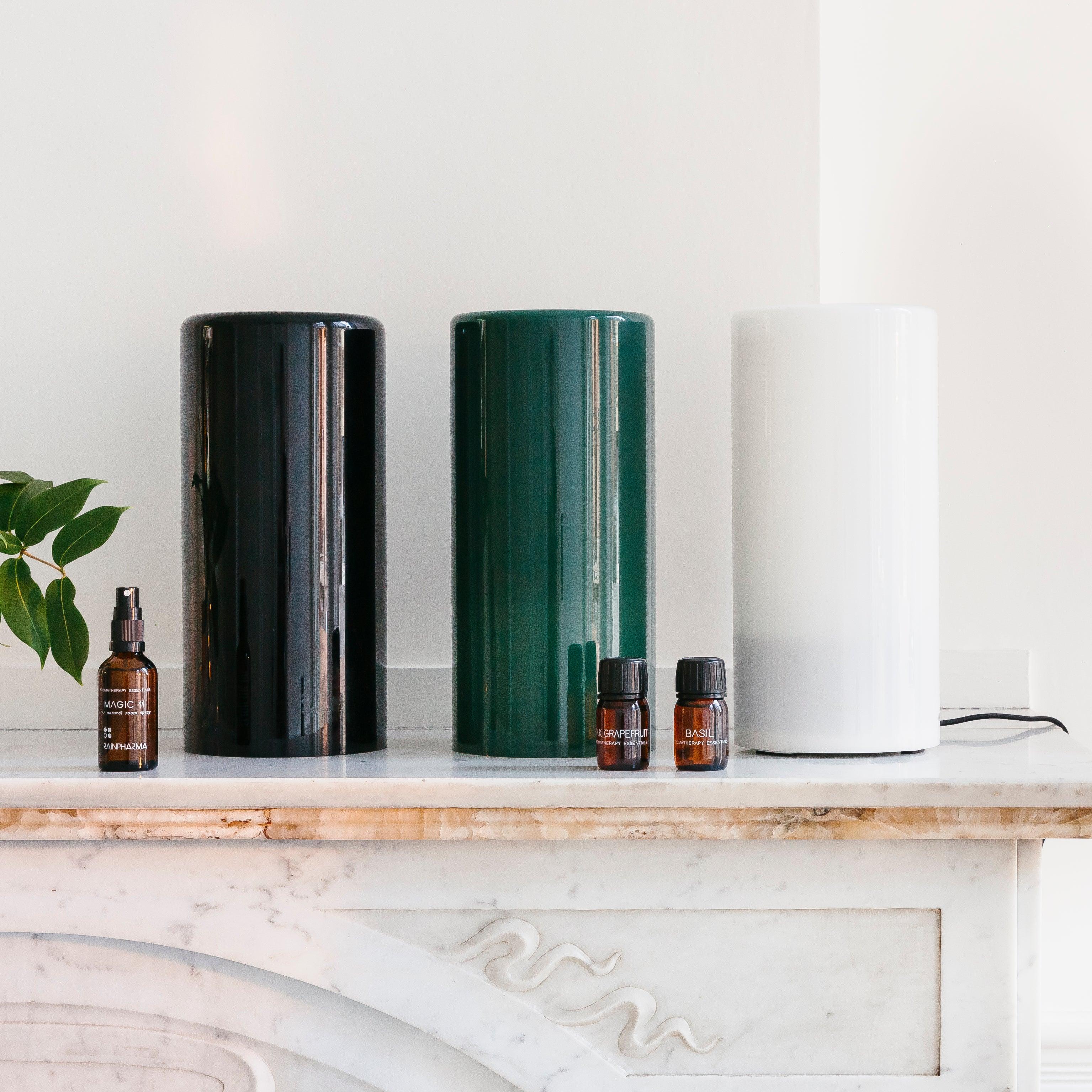Drie Aurora Aroma Diffusers van RainPharma, met glazen hulzen in de kleuren zwart, groen en wit, staan op een marmeren schouw naast kleine amberkleurige flesjes voor geurverspreiding en sfeervol licht.