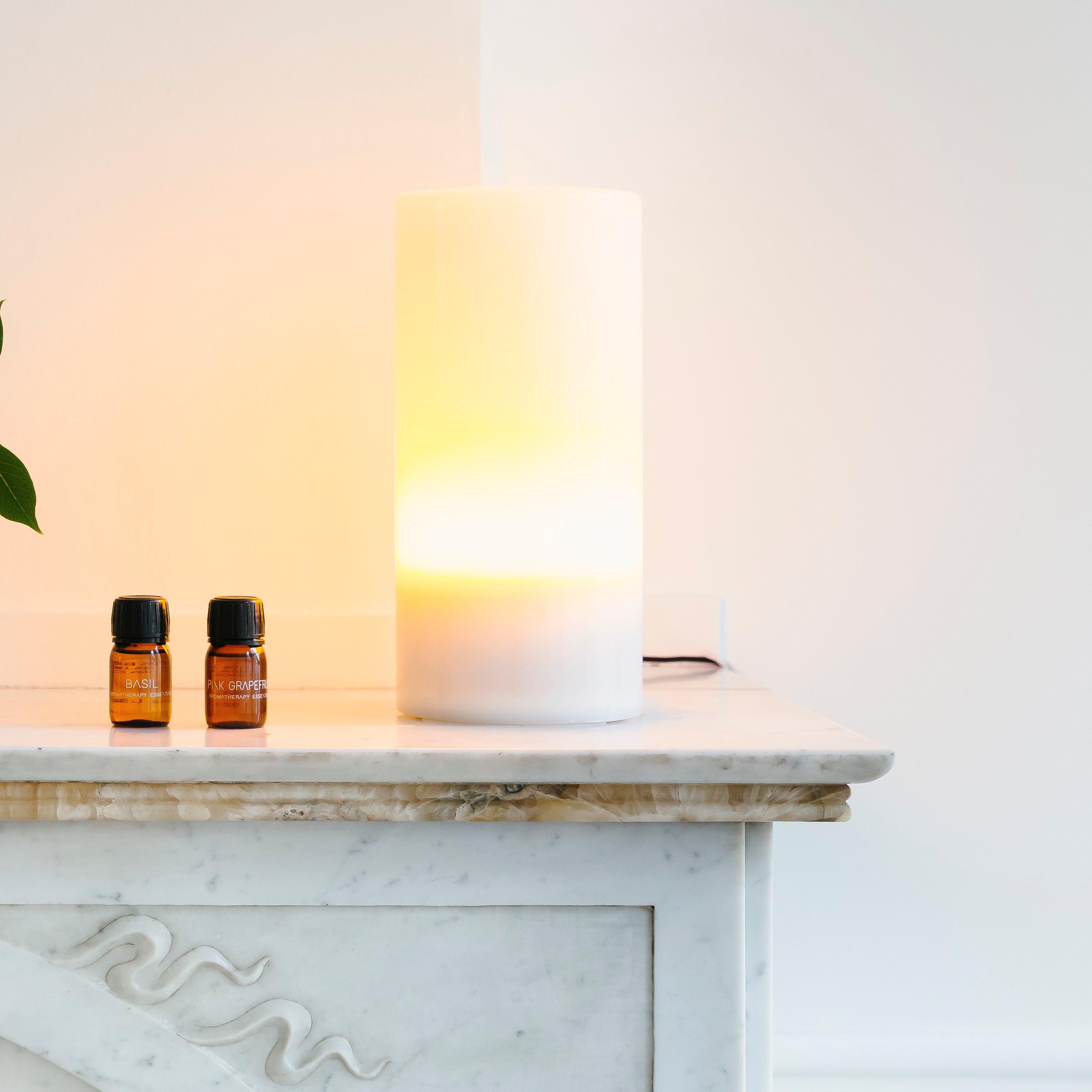 Een witte cilindervormige Aroma Diffuser - Aurora Glass Sleeve (kies uw kleur) van RainPharma en twee kleine amberkleurige flesjes staan op een marmeren oppervlak tegen een effen muur en werpen sfeervol licht door de kamer.