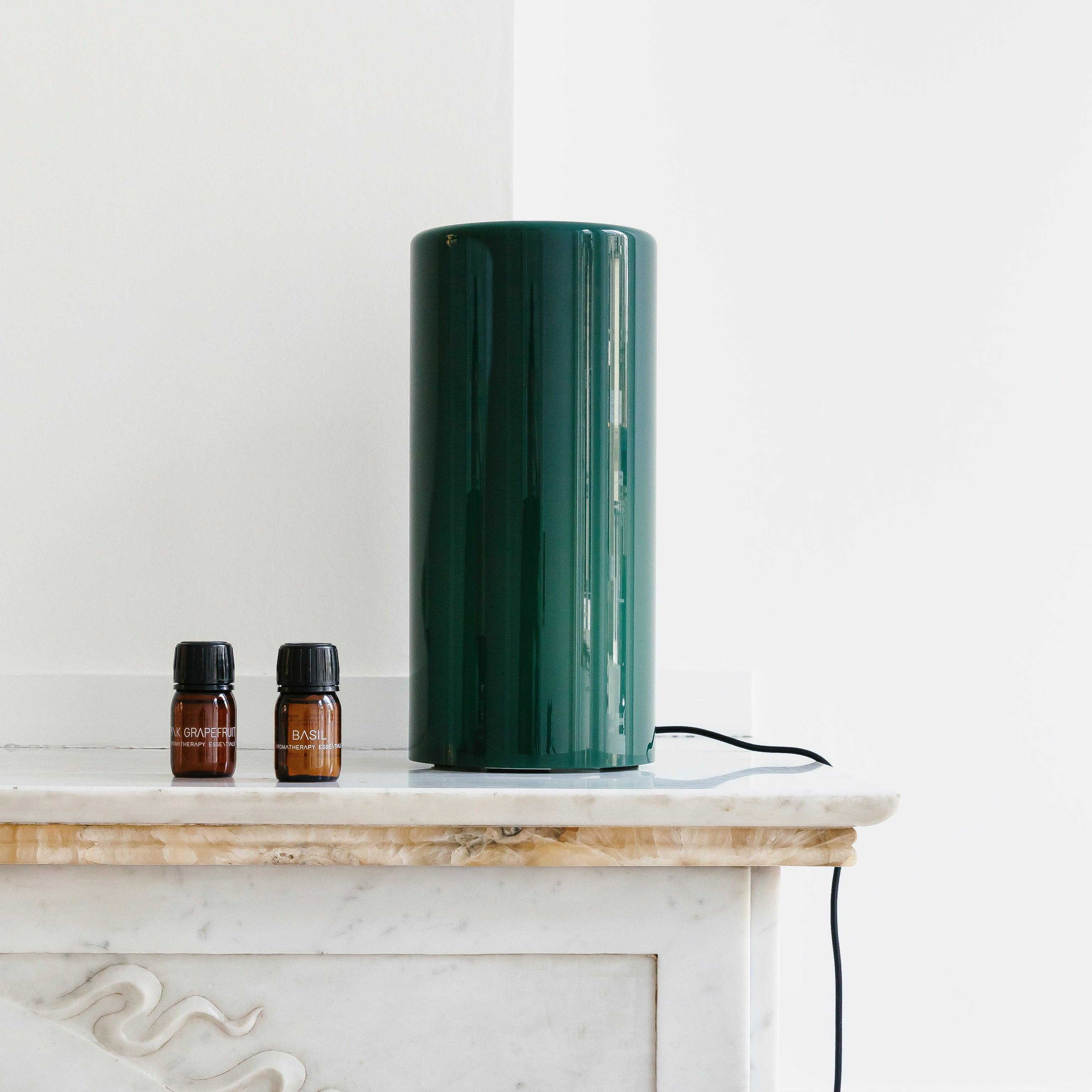 Een hoge, groene RainPharma Aroma Diffuser met een Aurora Glass Sleeve werpt sfeervol licht op de marmeren schoorsteenmantel naast twee kleine bruine flesjes etherische olie.