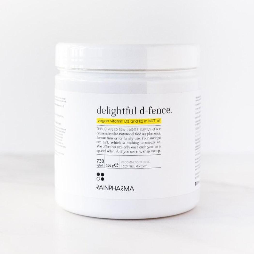 Een witte RainPharma-verpakking met het label "Vitamine D (Delightful D-fence)" bevat een veganistisch vitamine D- en K2-supplement in MCT-olie ter ondersteuning van weerstand, met productinformatie en geel uitgelichte tekst op de voorkant.