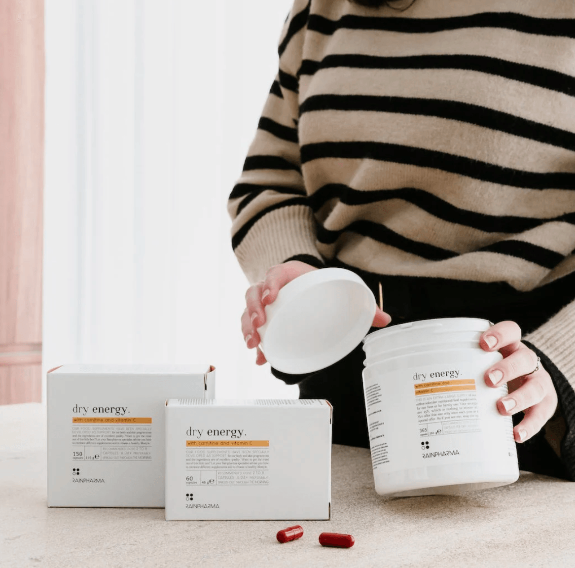 Een persoon in een gestreepte trui houdt een potje "Dry Energy - Family Pack 365 caps" van RainPharma vast, met dozen en rode capsules verspreid op tafel, die de vetstofwisseling bevorderen.