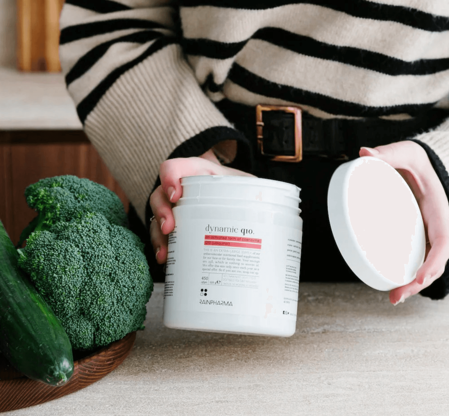 Een persoon opent een potje Dynamic Q10 - Family pack 450 caps van RainPharma, een krachtig voedingssupplement, naast knapperige komkommer en verse broccoli op het aanrecht.
