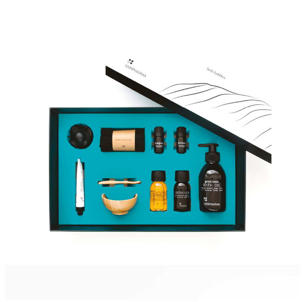 Open doos van Luxe Bad Set - Bath bubbles van RainPharma, met badproducten zoals flessen, een houten schep, borstel en een tube etherische olie. Gerangschikt op een blauwe achtergrond met het gedeeltelijk open deksel dat een minimalistisch ontwerp onthult. Ideaal om uw badritueel om te vormen tot een luxe thuisspa.