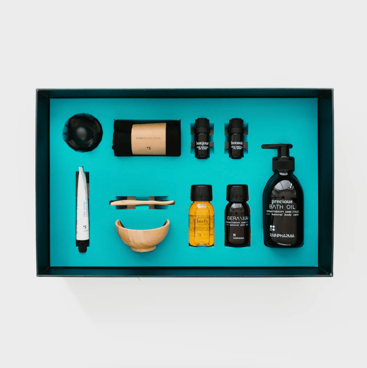 De Luxe Bad Set van RainPharma, op een turquoise achtergrond, bevat badolie, handcrème, twee essentiële oliën, een houten kom, druppelaar en borstel voor de perfecte spa-ervaring bij u thuis.