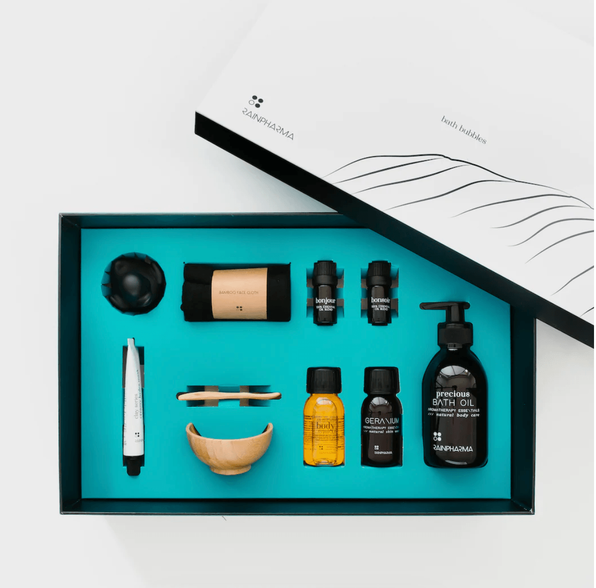 Open Luxe Bad Set van RainPharma op een turquoise achtergrond, met essentiële olie, zeep en een houten kom. Het gedeeltelijk getoonde deksel heeft een lijnkunst-branding, perfect om uw badritueel om te vormen tot een rustige thuisspa-ervaring.