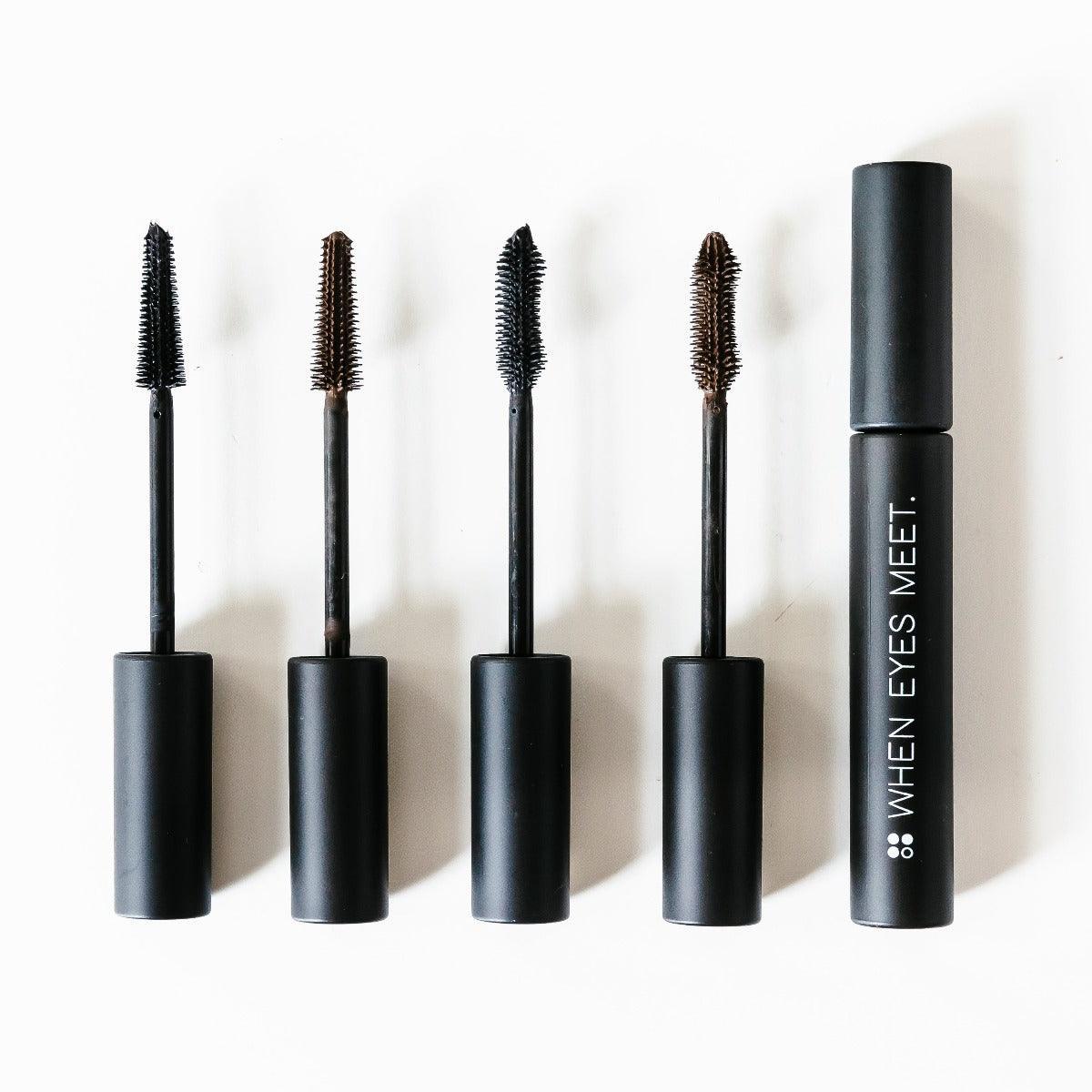Vier mascaraborstels met verschillende borstelharen worden getoond bij een tube met het label "When eyes meet" op een witte achtergrond. Deze RainPharma veganistische mascara in natuurlijk bruin gebruikt cannabis sativa-olie voor opvallende, langdurige wimpers.