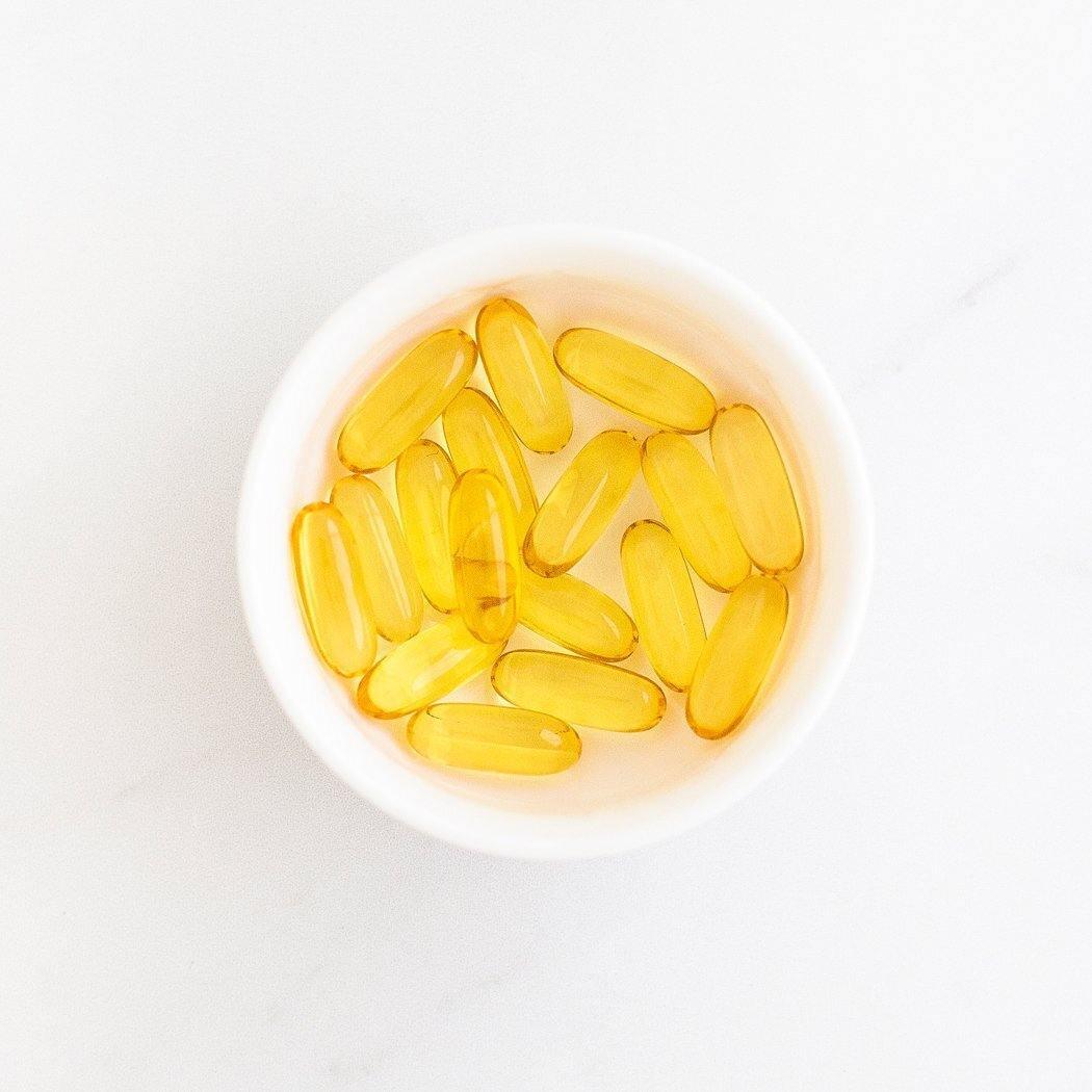 Een witte kom bevat verschillende gele, ovaalvormige Ultra Omega 3 softgel capsules van RainPharma, rijk aan EPA en DHA omega-3 vetzuren, die rusten op een wit oppervlak.