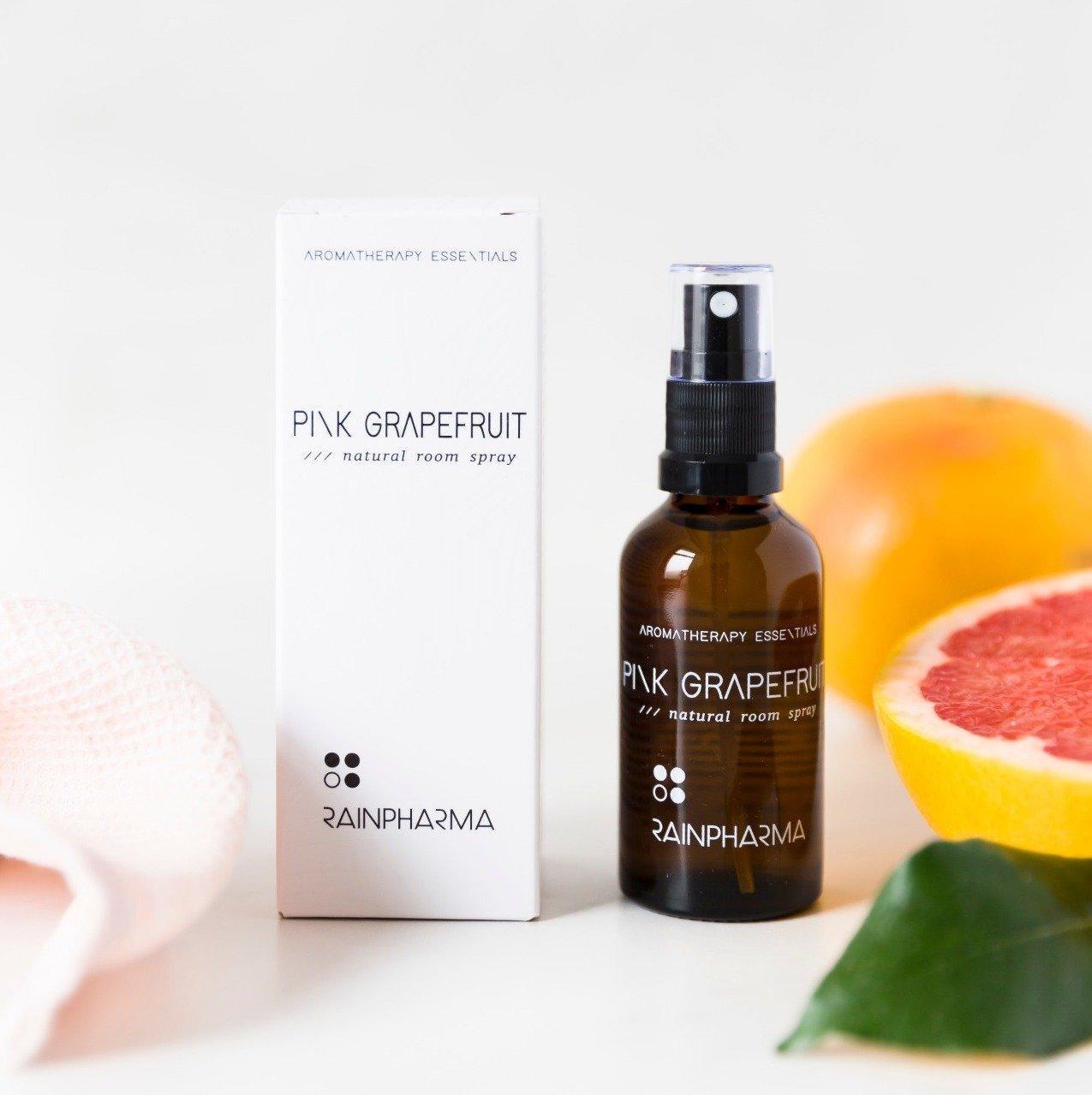 Een RainPharma Room Spray Pink Grapefruit fles en doosje staan naast een gehalveerde grapefruit, een blad en een roze doek op een wit oppervlak.