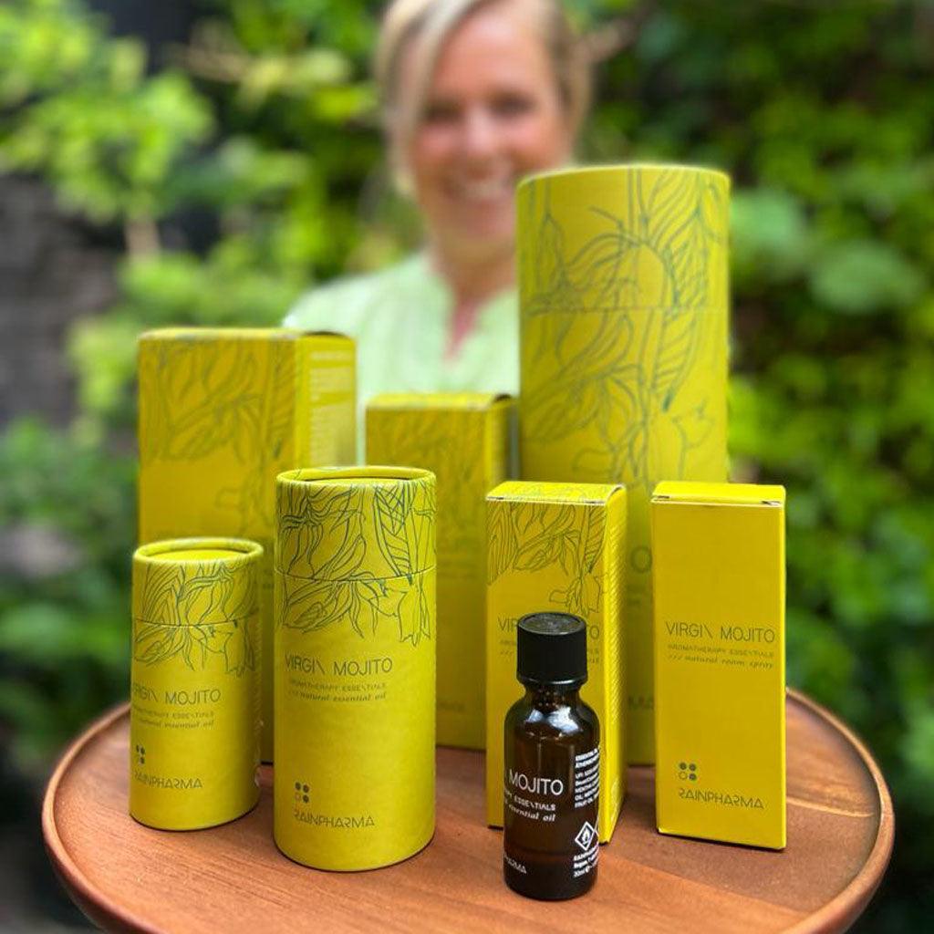 Gele doosjes met RainPharma’s Natural Room Spray - Virgin Mojito en een klein donker spuitflesje staan op een houten tafel buiten, met een wazig persoon zichtbaar op de achtergrond.