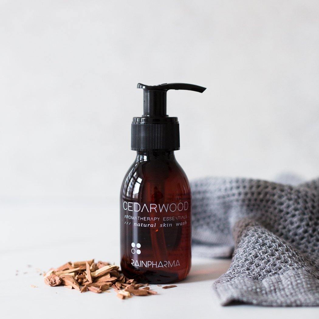 Een bruine pompflacon van RainPharma Skin Wash Cedarwood, gemaakt met natuurlijke ingrediënten, staat naast cederhoutkrullen en een grijze textuurdoek op een wit oppervlak.