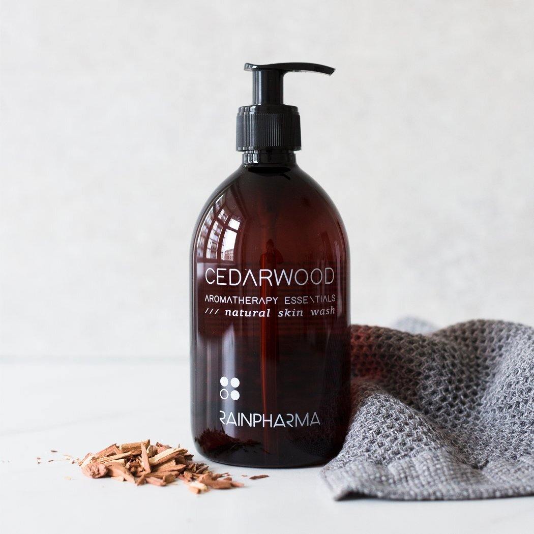 Een fles RainPharma Skin Wash Cedarwood met pompje, naast een grijze handdoek en houtsnippers op een licht oppervlak, doordrenkt je dagelijkse routine met de kalmerende cedergeur en natuurlijke ingrediënten.