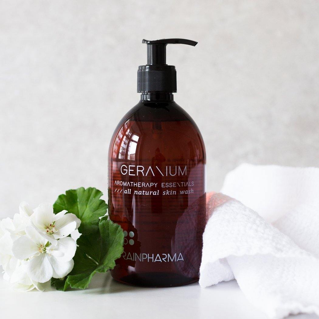 Naast een witte handdoek en witte bloemen met groene bladeren staat een bruine pompflacon met de tekst "RainPharma Skin Wash Geranium".