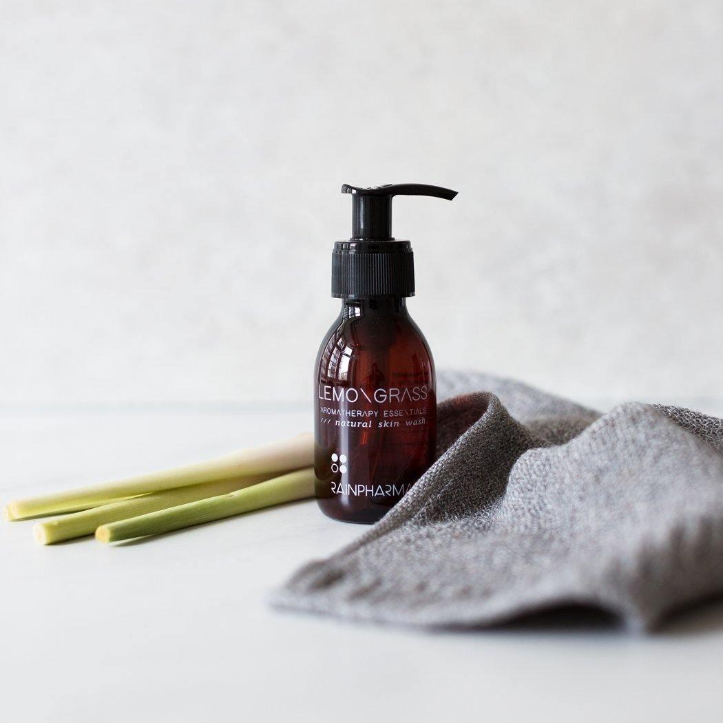 Een bruine pompflacon van RainPharma Skin Wash Lemongrass rust op een oppervlak naast verse citroengrasstengels en een gevouwen grijs doekje, wat de natuurlijke inspiratie van deze douchegel op basis van planten weerspiegelt.