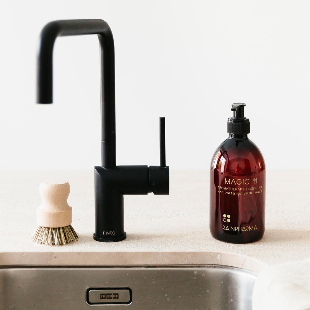 Een keukenopstelling met een strakke zwarte kraan, een houten schrobber en een fles RainPharma Skin Wash Magic 11 met natuurlijke ingrediënten op het aanrecht.
