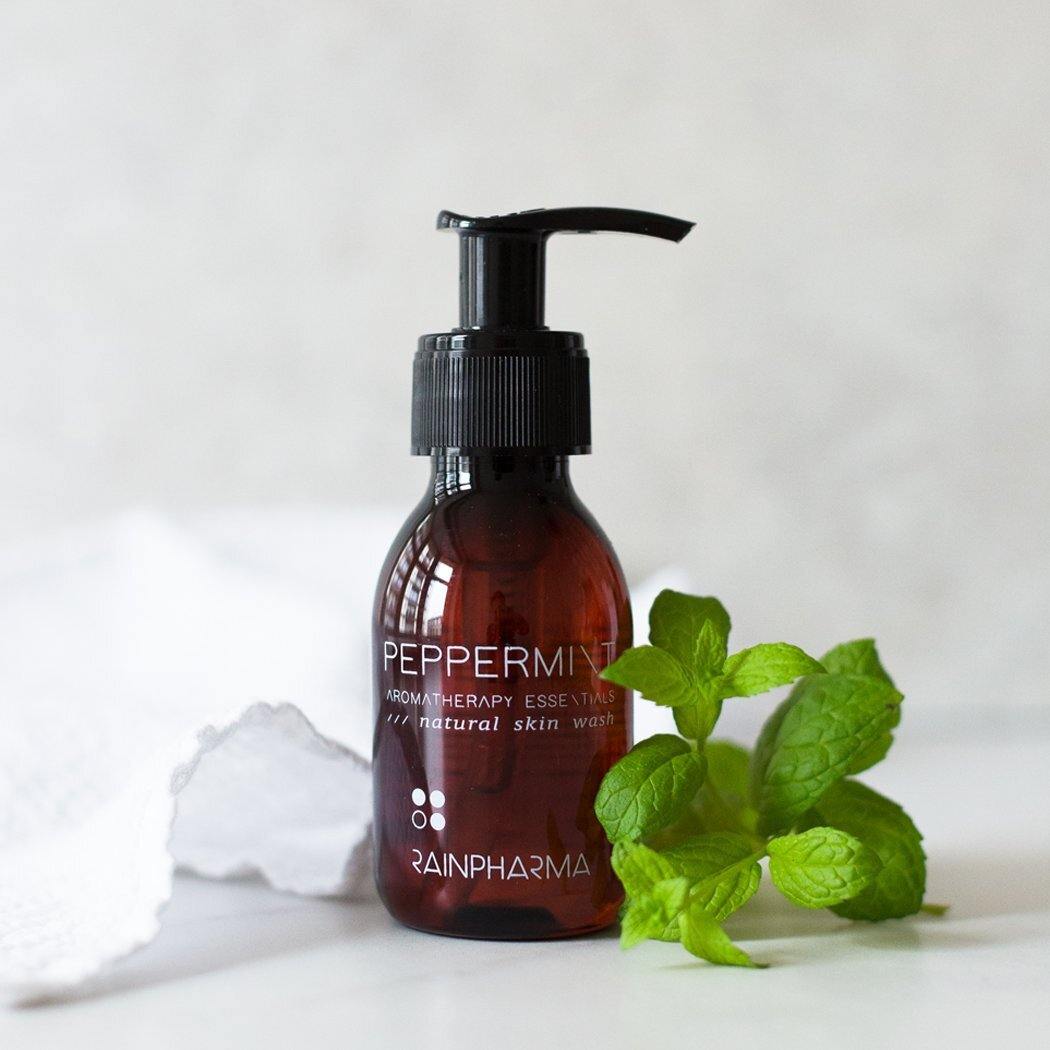 Een bruine pompflacon RainPharma Skin Wash Peppermint, een plantaardige douchegel, staat op een wit oppervlak naast een vers munttakje en een witte handdoek.