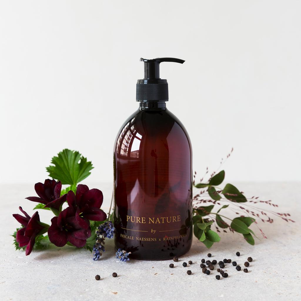 Een bruine pompflacon van Skin Wash Pure Nature van Pascale Naessens van RainPharma rust op een licht oppervlak, omringd door donkere bloemen, groen en verspreide peperkorrels.