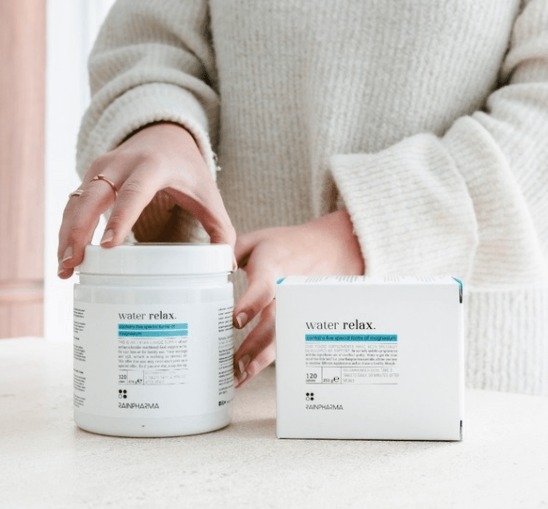 Een persoon in een trui raakt een pot aan met het opschrift "Water Relax - Familieverpakking 320 caps" van RainPharma, verrijkt met magnesium, naast een bijpassend doosje op het aanrecht.