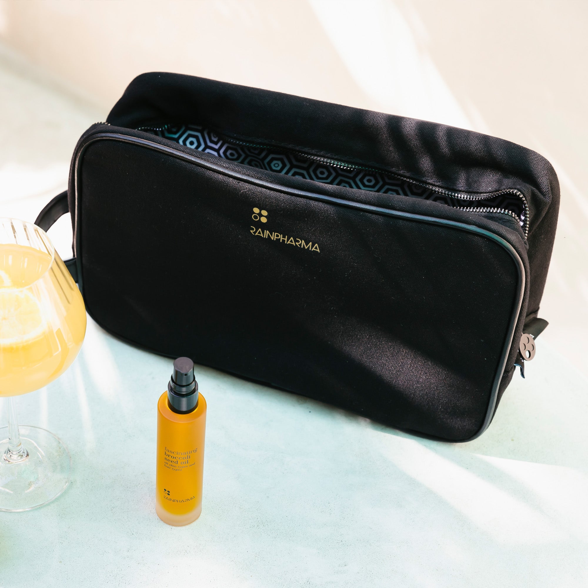 De RainPharma Toilettas - Maxi - Zwart staat op een lichte ondergrond naast een glas sinaasappelsap en een gele fles met pompje. Ideaal voor op reis of om je verzorgingsproducten in op te bergen.