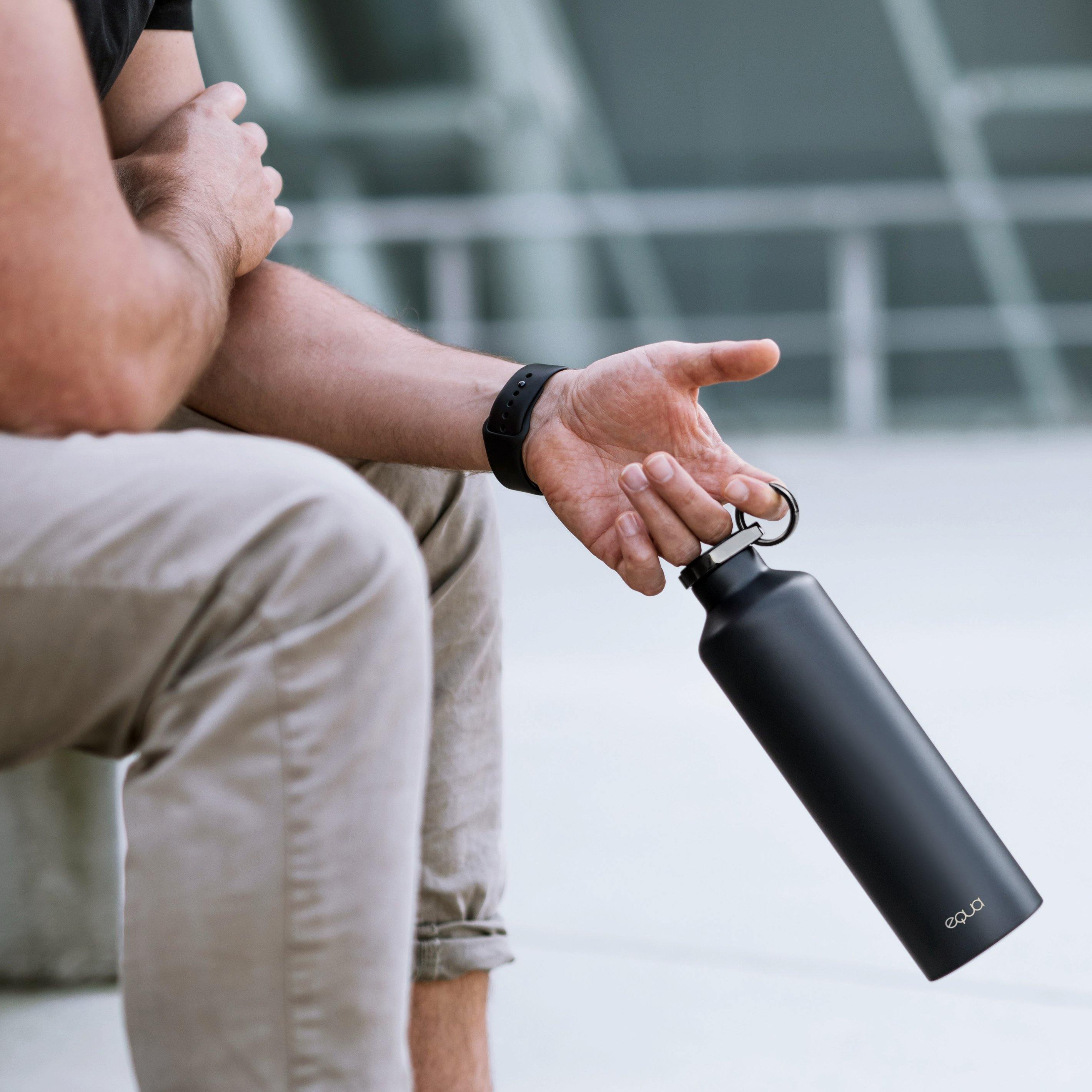 Buiten houdt iemand de Equa Waterfles Thermo Stainless steel in Dark Grey vast aan de lus, de arm rust op de knie. Deze BPA-vrije fles van Equa is gemaakt van roestvrij staal - ideaal voor duurzame hydratatie.
