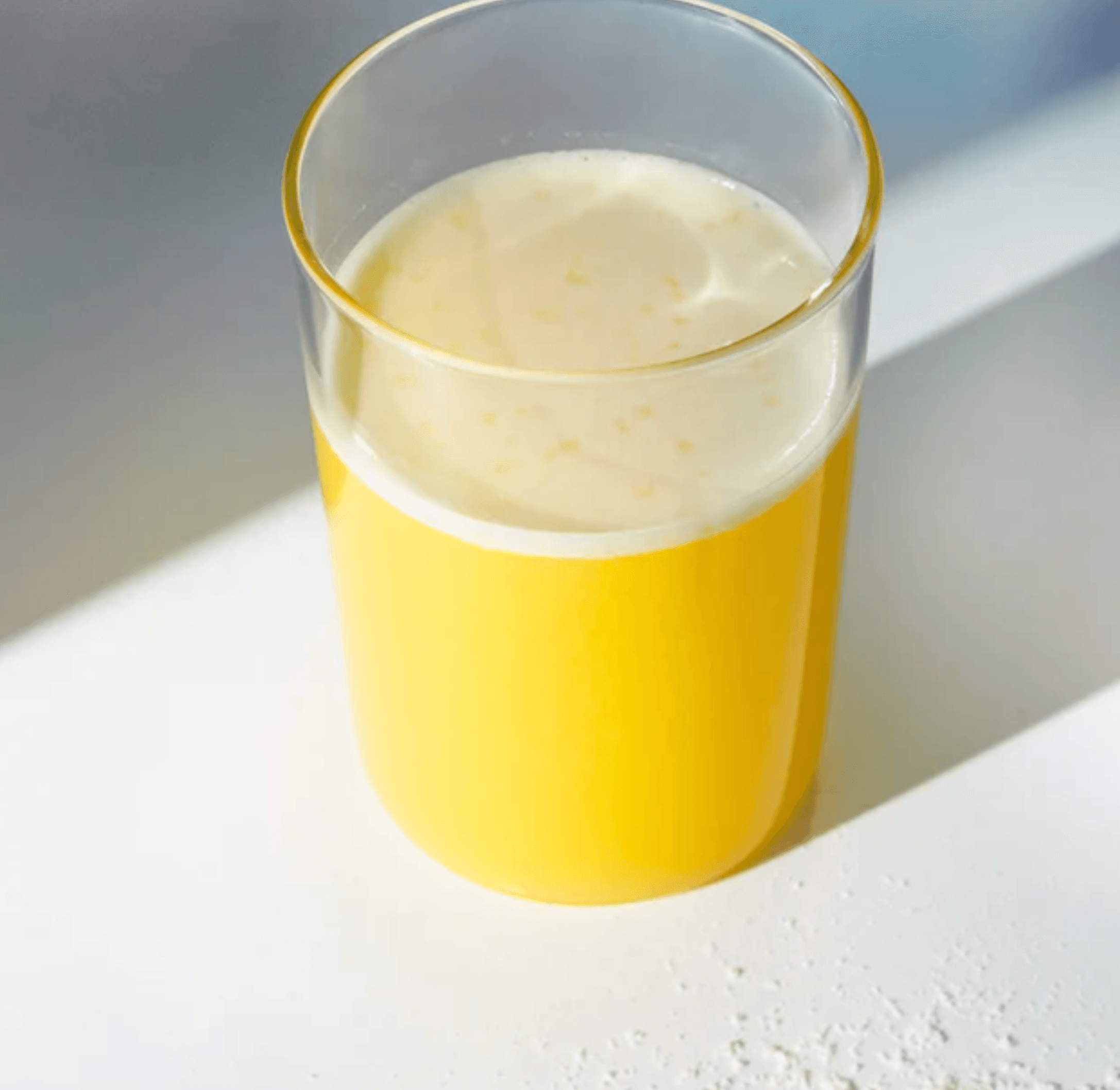 Een glas geel-sinaasappelsap, verrijkt met eiwitten, staat op een wit oppervlak en werpt een schaduw. Deze verfrissende drank is van RainPharma&