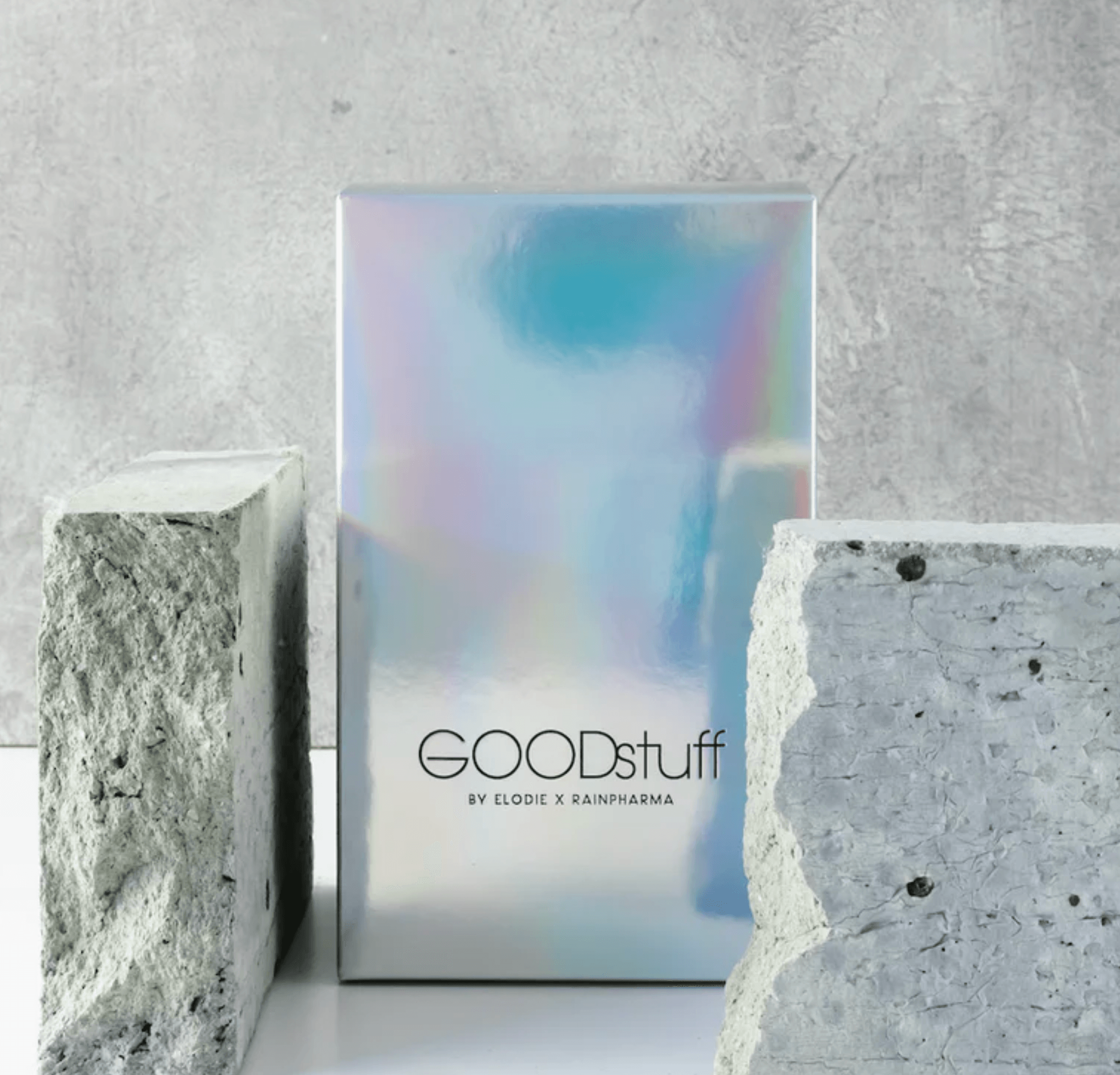 Een glimmende holografische doos met het opschrift "1 zakje GOODstuff by Elodie x RainPharma", bekend om zijn hoogwaardige eiwitten voedingssupplement, is tussen twee grote betonblokken op een grijze achtergrond geplaatst.