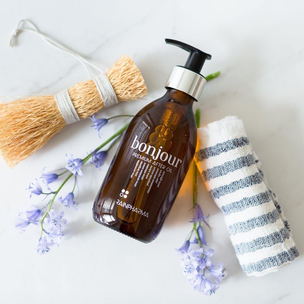 Een flesje van 250 ml RainPharma Premium After Oil - Bonjour (Deluxe), door aromatherapeuten gewaardeerd om zijn hoogwaardige essentiële oliën, ligt op een wit oppervlak met een gestreepte handdoek, een natuurlijk kwastje en paarse bloemen.