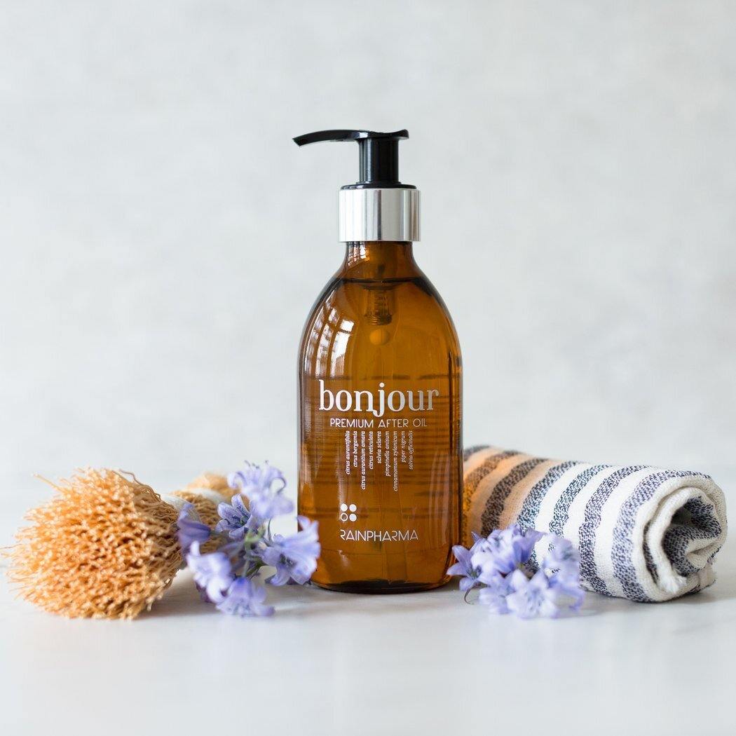 Een amberkleurig flesje Bonjour Premium After Oil van RainPharma, verrijkt met essentiële oliën, wordt omringd door paarse bloemen, een natuurlijke spons en een opgerolde gestreepte handdoek op een licht oppervlak.