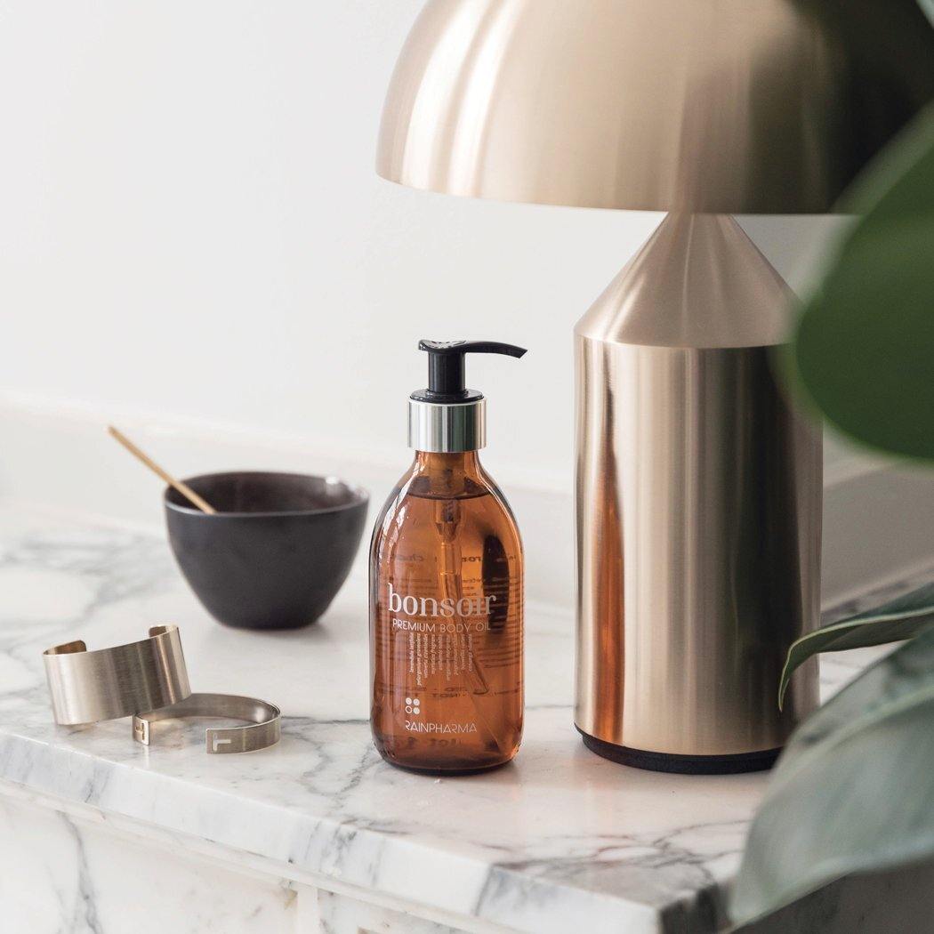 Een pompflacon Bonsoir Premium Body Oil van RainPharma staat elegant op een marmeren oppervlak, naast een moderne messing lamp, een klein kommetje met een lepel en strakke metalen accessoires.