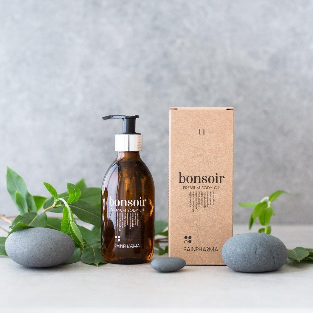 Naast de doos is een flesje RainPharma Premium Body Oil - Bonsoir (Deluxe) van 250 ml met pompje te zien, omringd door gladde grijze stenen en groene bladeren, die een aromatherapeutische geur oproepen.