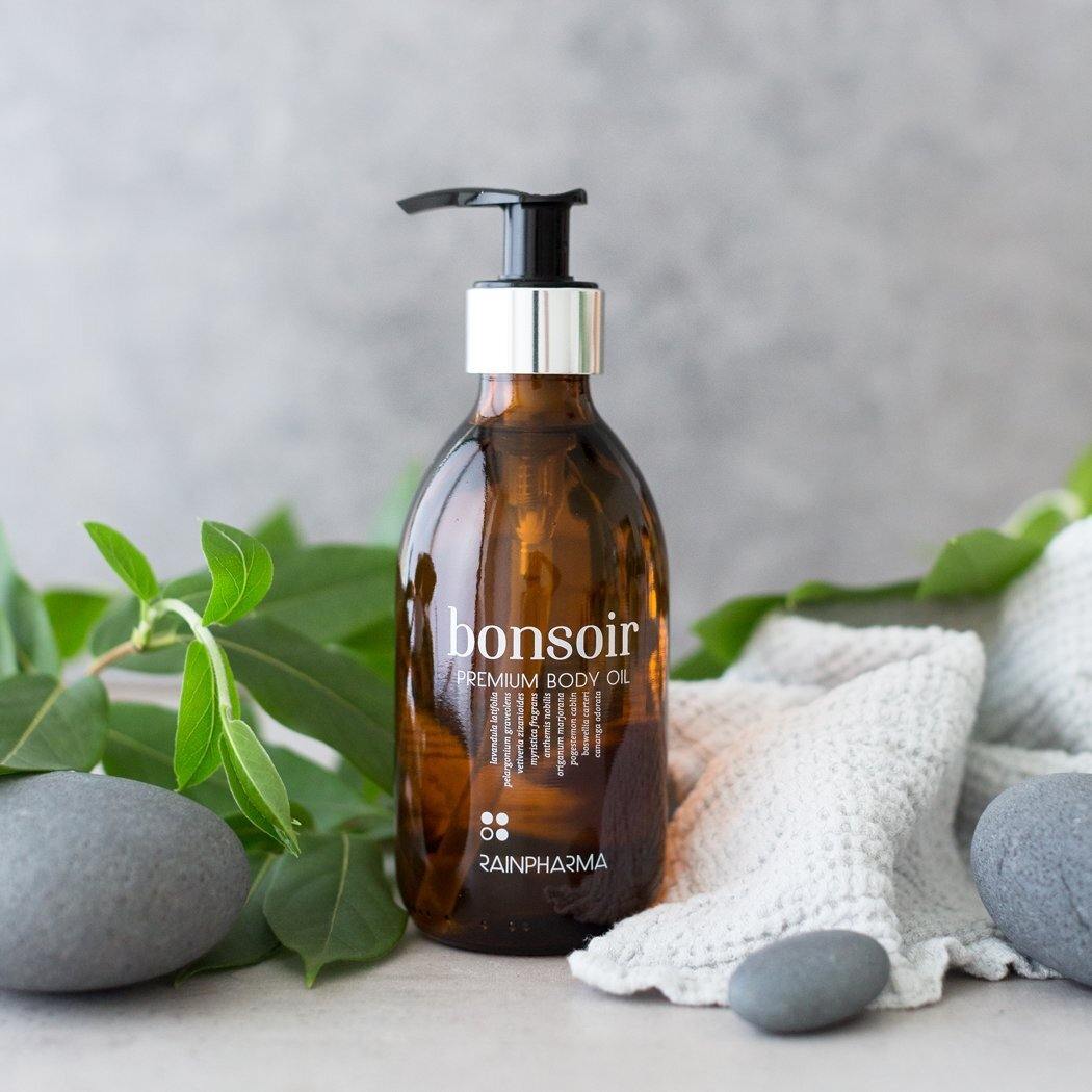 De amberkleurige fles Bonsoir Premium Body Oil van RainPharma heeft een pompdispenser en is elegant omgeven door groene bladeren, grijze kiezels en een grijze stof met structuur.