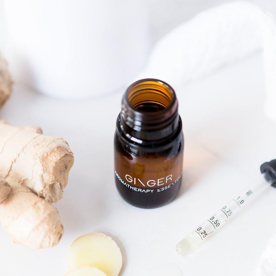 Een klein amberkleurig flesje met het label "Essentiële olie - Gember" van RainPharma wordt tentoongesteld met gemberwortel, gesneden gember en een druppelaar op een onberispelijk wit oppervlak, wat de krachtige therapeutische eigenschappen ervan benadrukt.