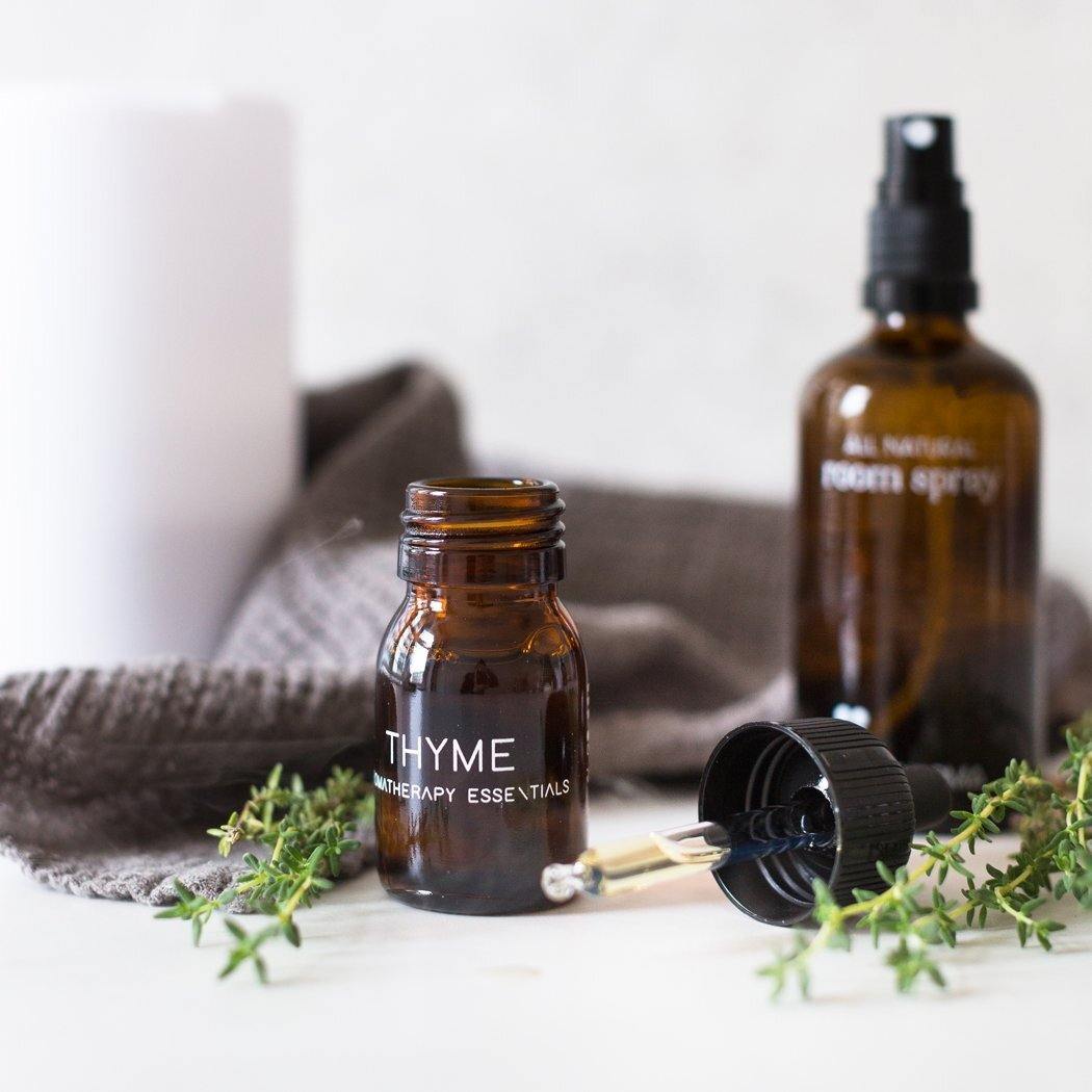 De amberkleurige flesjes etherische olie tijm van RainPharma, met de labels "Tijm" en "Kamerspray", zijn voorzien van een druppelaar en zijn op een witte ondergrond op kunstzinnige wijze omringd door takjes tijm, aangevuld met een grijze doek die verwijst naar de rijke therapeutische eigenschappen van de olie.