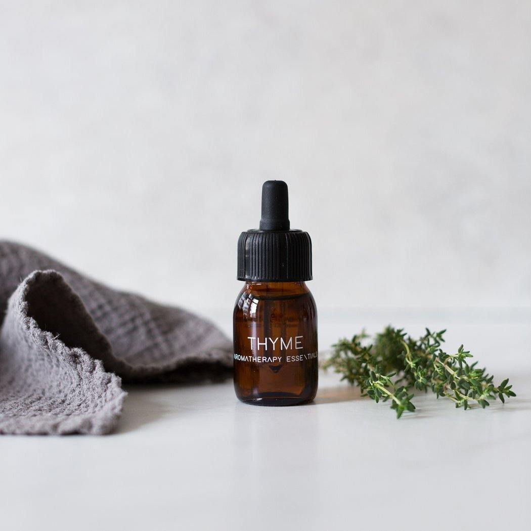 Een klein amberkleurig flesje "RainPharma Essential Oil Thyme" staat naast verse tijm en een grijze doek op een licht oppervlak, wat de therapeutische eigenschappen van deze krachtige etherische olie belichaamt.
