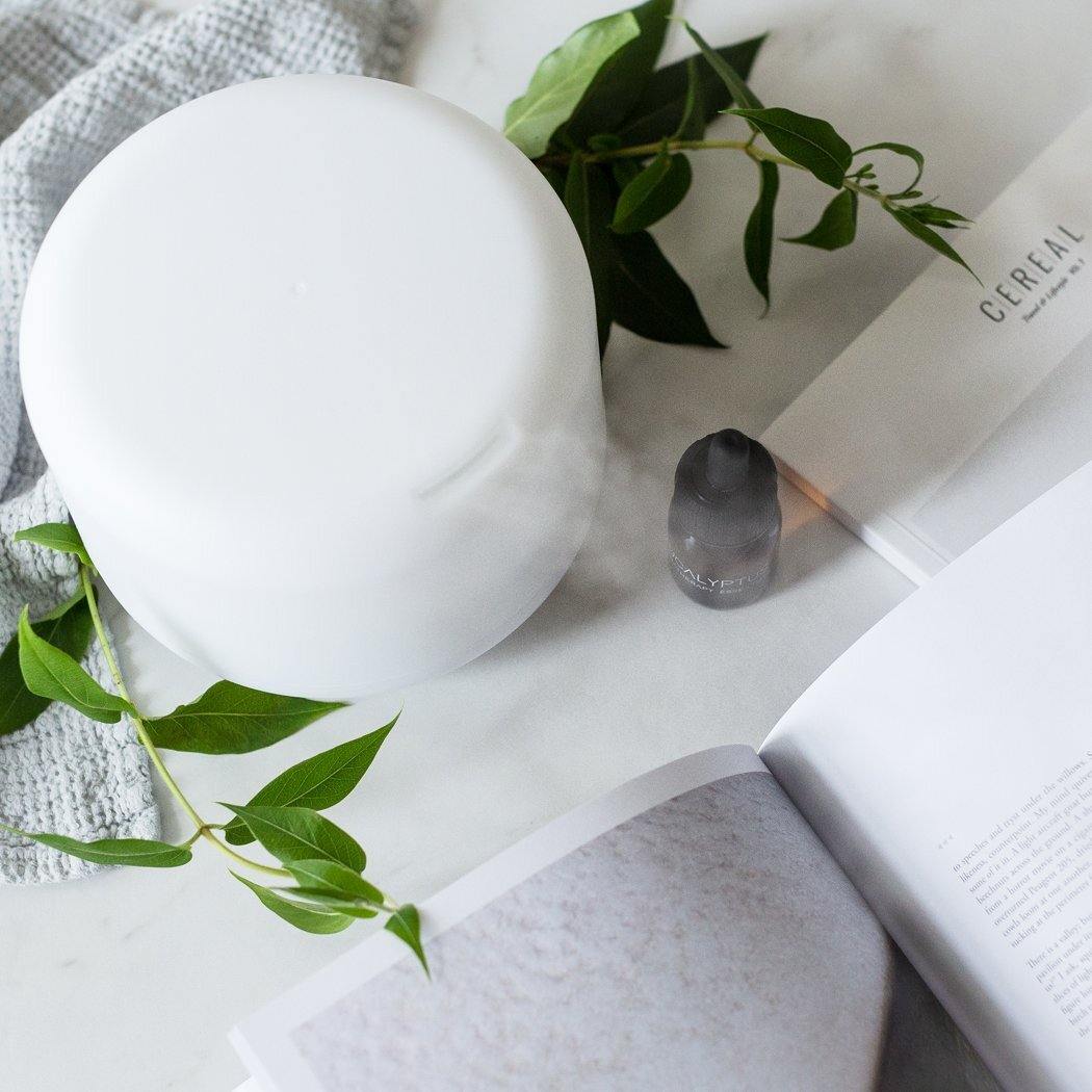 Een RainPharma Aroma Diffuser staat naast een klein olieflesje, groene bladeren, een handdoek en een opengeslagen tijdschrift op een wit oppervlak.