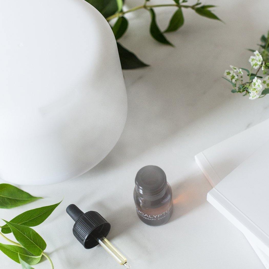Een klein druppelflesje met een losse pipet staat op een wit oppervlak naast de RainPharma Aroma Diffuser van RainPharma, ideaal voor het verspreiden van etherische oliën, samen met bladertakken en een wit notitieblok.