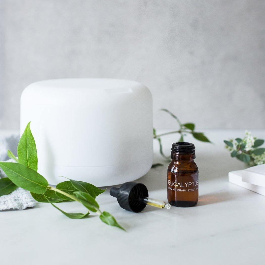 De RainPharma Aroma Diffuser staat op een wit oppervlak naast een klein bruin eucalyptusflesje, druppelaar en groene bladeren en verspreidt met behulp van ultrasone golven op zachte wijze rustgevende aroma&