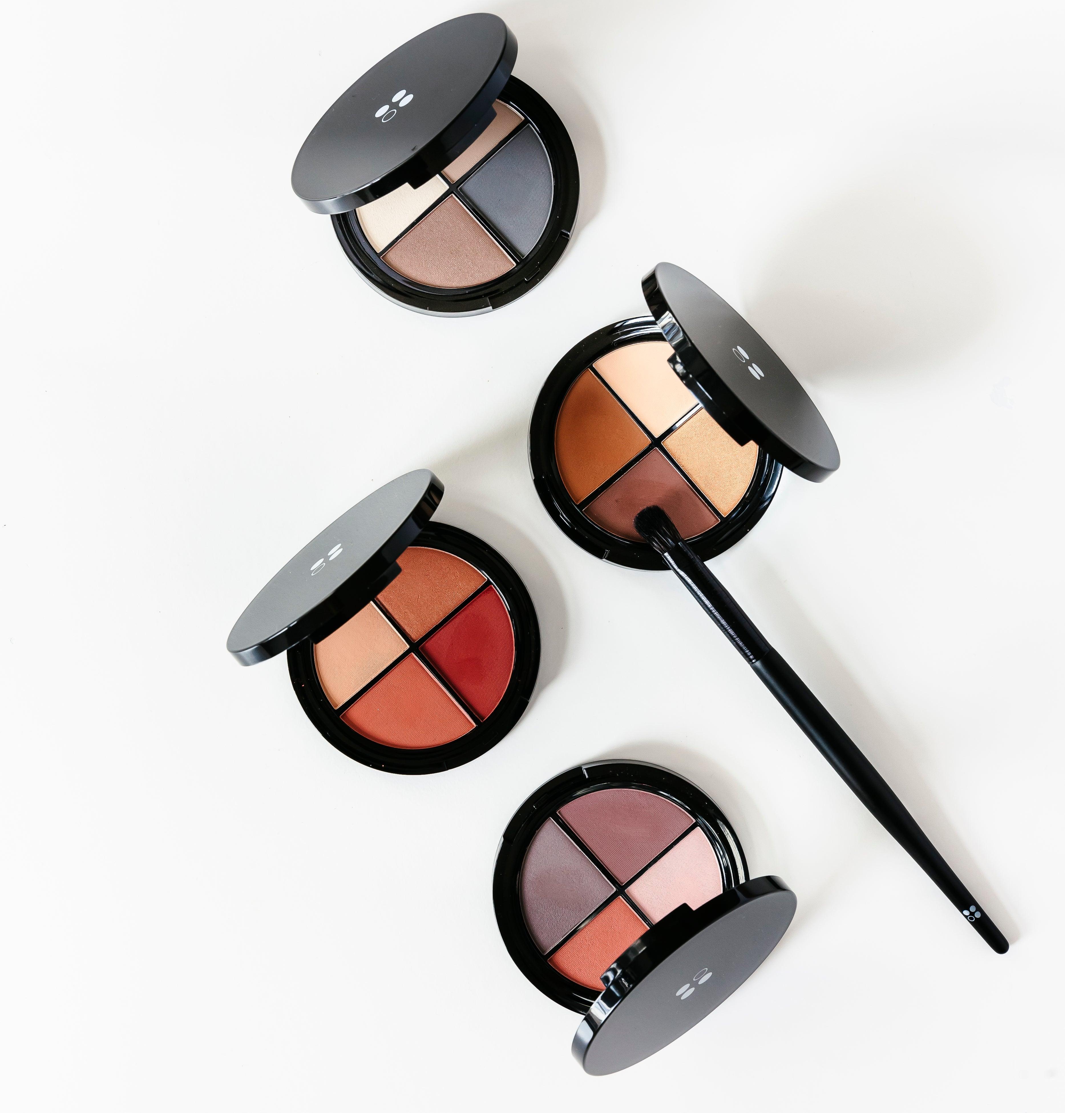 Vier ronde oogschaduwpaletten van Puur Living, geïnspireerd op Cadeaubon Make-up verwenmoment voor twee, liggen op een wit oppervlak - één met een make-upkwast erop.