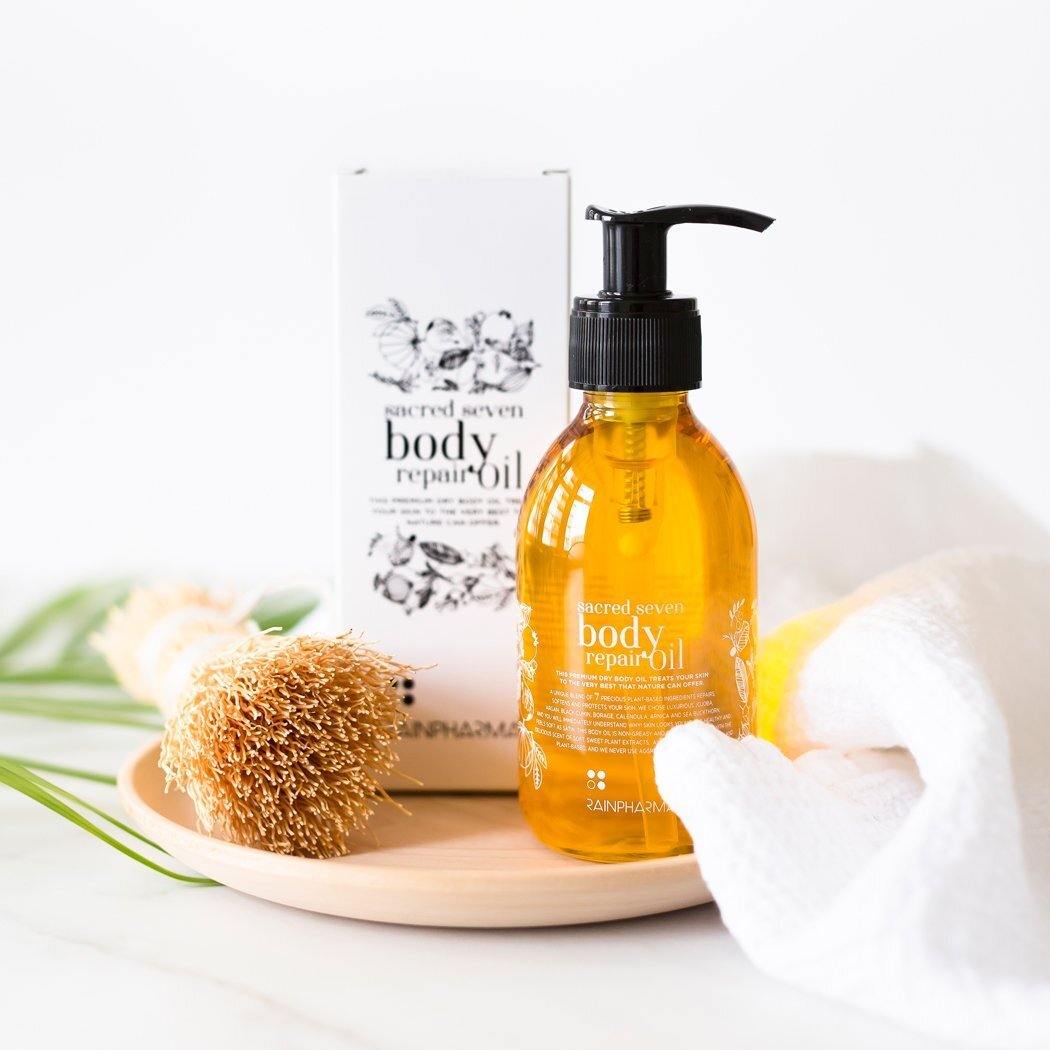 De Sacred Seven Body Repair Oil van RainPharma, gemaakt van plantaardige ingrediënten, ligt op een houten bord naast een witte handdoek en een borstel met structuur. De verpakking is op de achtergrond zichtbaar.