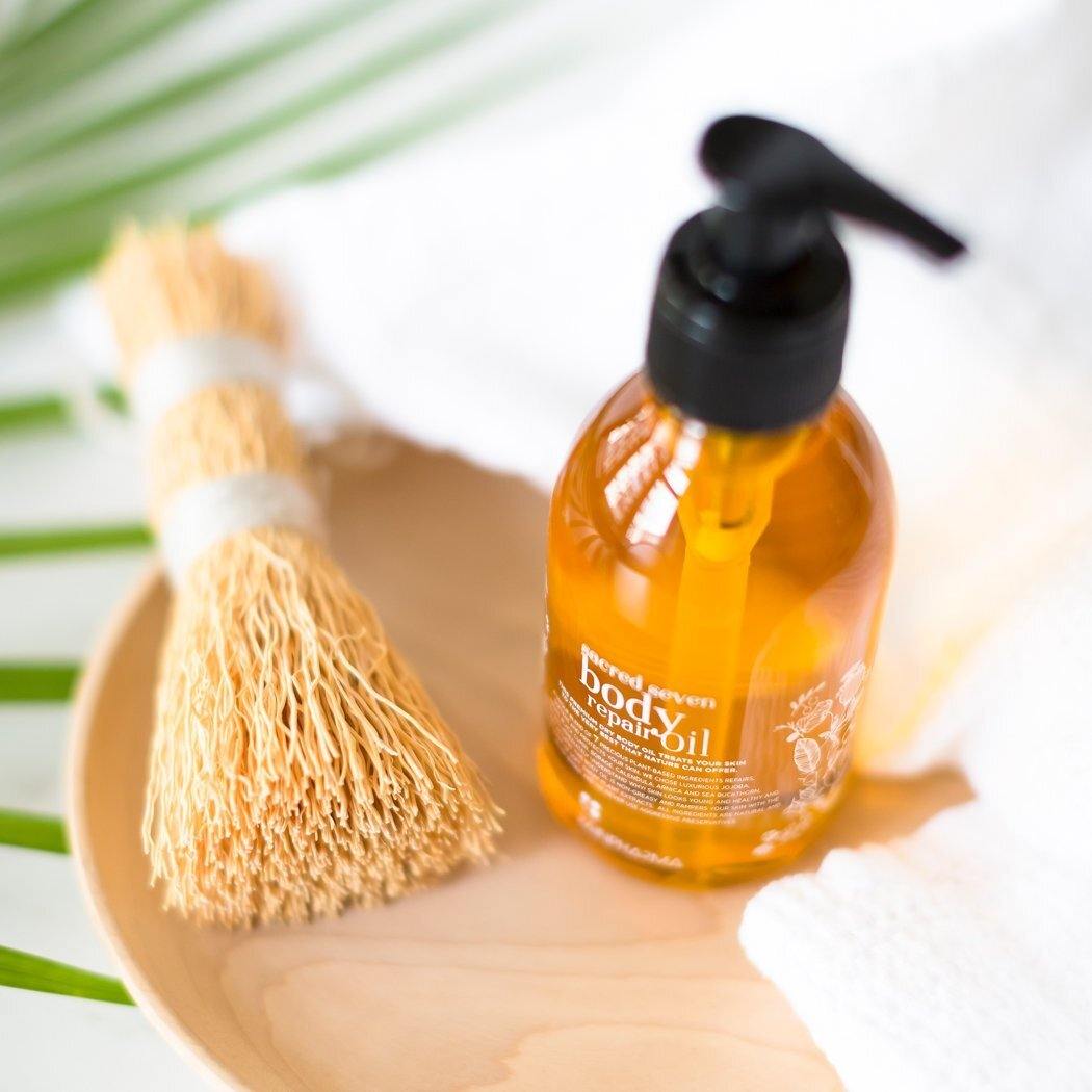 Een flesje Sacred Seven Body Repair Oil van RainPharma, voorzien van een zwart pompje en gemaakt van plantaardige ingrediënten, staat op een houten bord naast gedroogd stro.