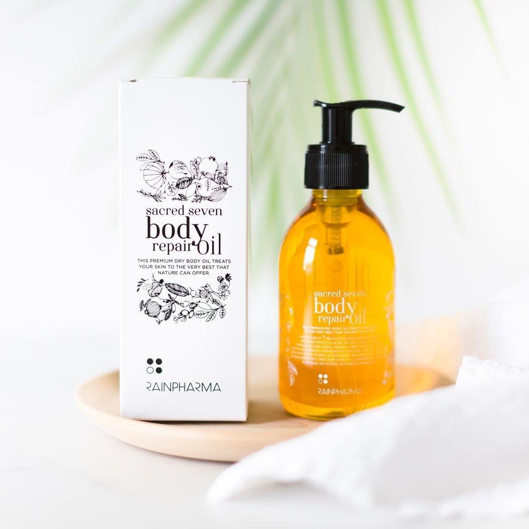 Een flesje Sacred Seven Body Repair Oil van RainPharma, rijk aan plantaardige ingrediënten, staat op een wit oppervlak naast de verpakking met bloemenmotief op een houten dienblad.
