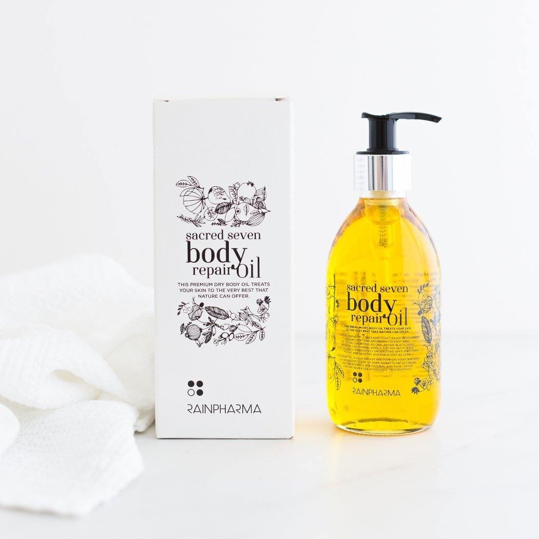 Een fles Sacred Seven Body Repair Oil van RainPharma, gemaakt met plantaardige ingrediënten, staat naast de doos, met een witte handdoek op de achtergrond.