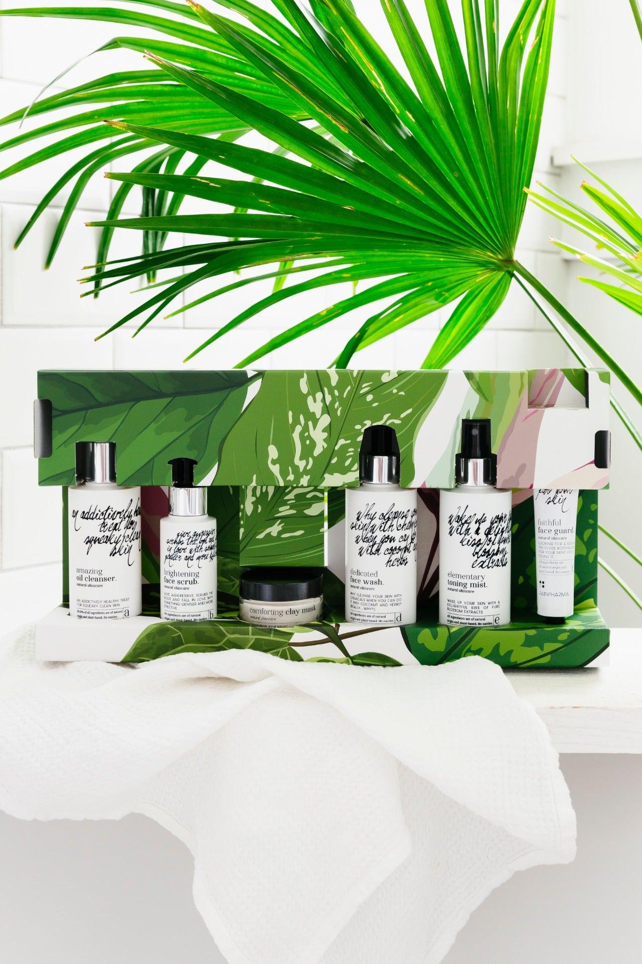 De Complete Skincare set - abcdef promobox 4+2 GRATIS van RainPharma heeft een elegante, natuurlijke groene en witte verpakking op een wit oppervlak, versterkt door een groot groen palmblad op de achtergrond, wat een holistische benadering van natuurlijke huidverzorging laat zien.