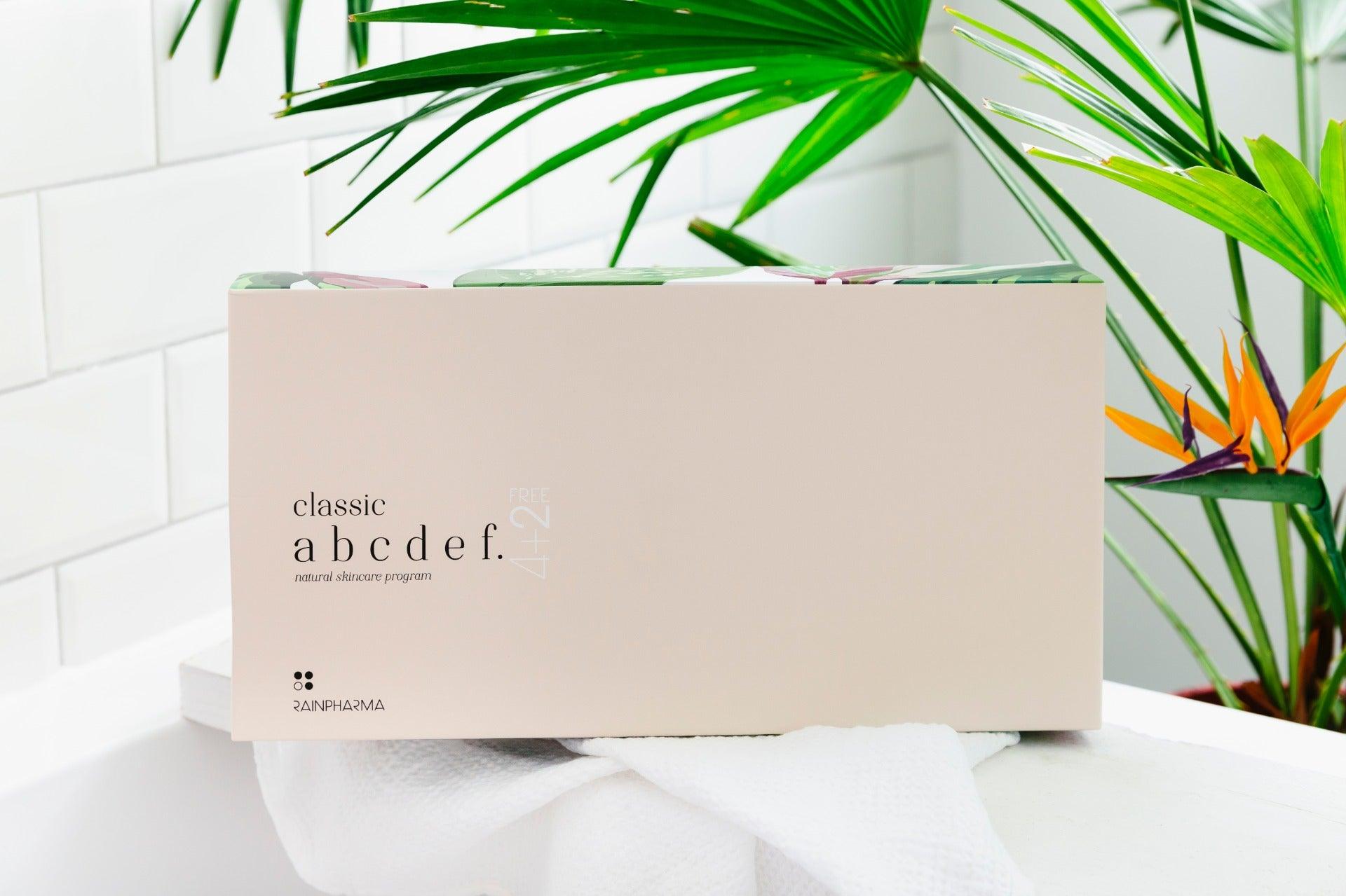 Een beige rechthoekige doos met de opschriften "Complete Skincare set - abcdef promobox 4+2 GRATIS" en "RainPharma" staat op een witte handdoek in een lichte badkamer met groene planten, wat staat voor een natuurlijke huidverzorgingsaanpak.
